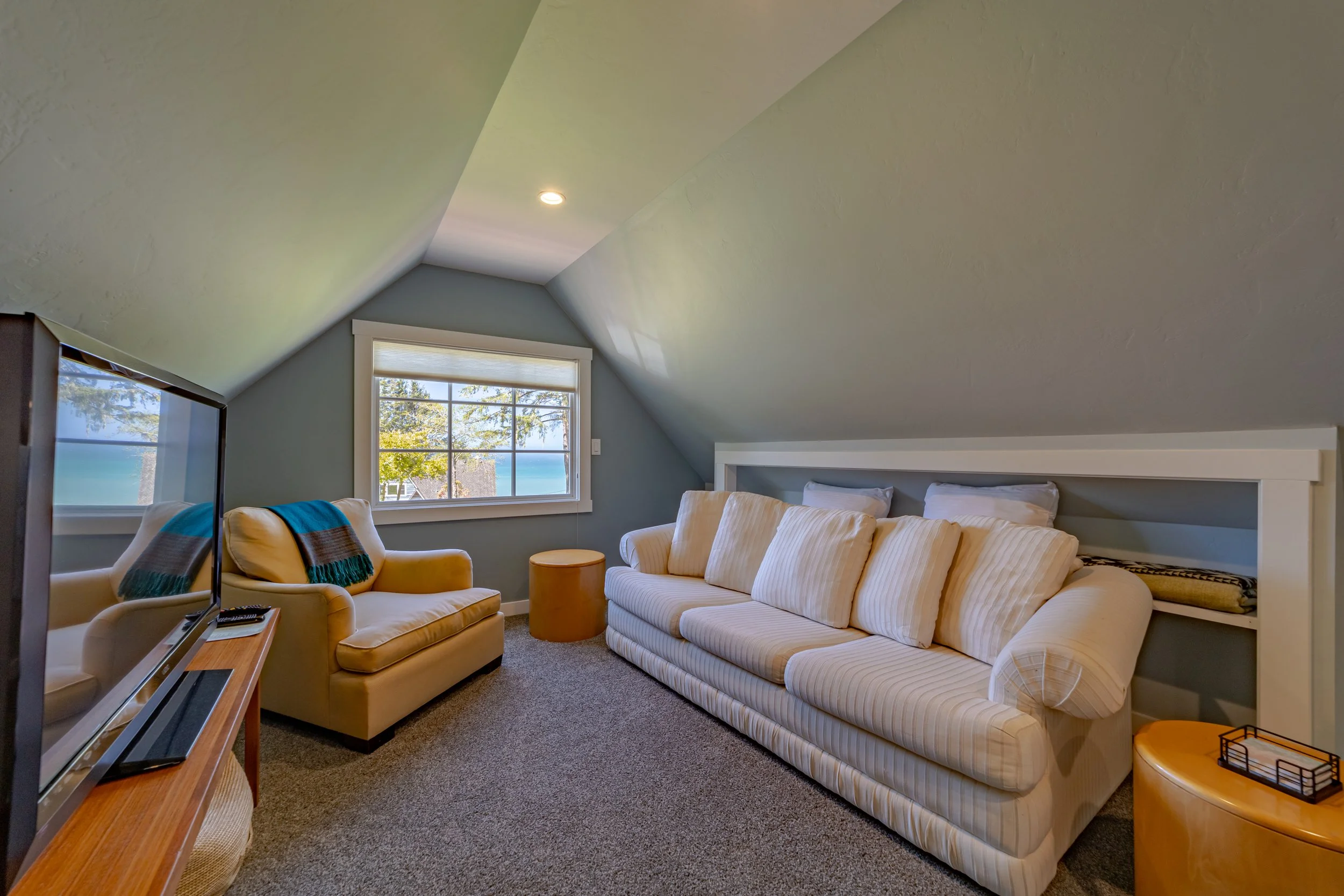 Cozy attic living room with sloped ceiling, featuring a large striped sofa, a beige armchair with a blue and brown throw, a window with a view of trees and water, a flat-screen TV on a wooden stand, and small round side tables.