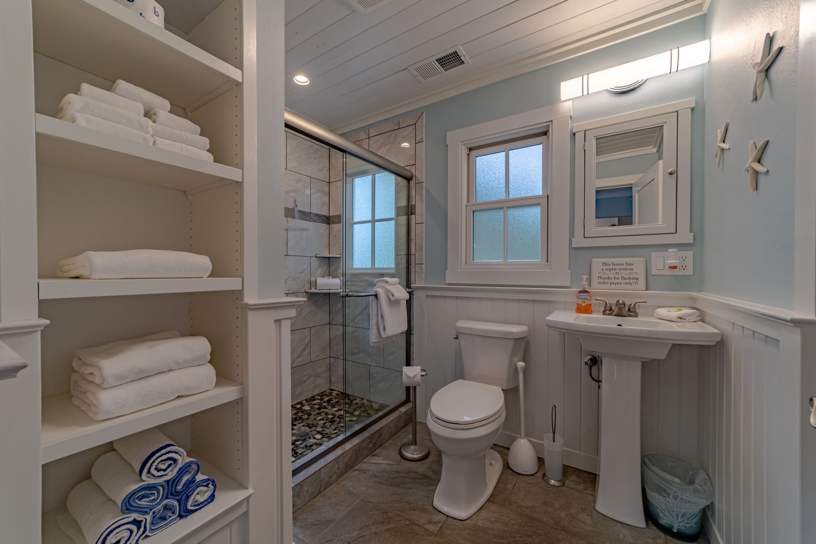 Bathroom with a shower, toilet, and small sink, decorated in light colors with storage shelves holding towels.