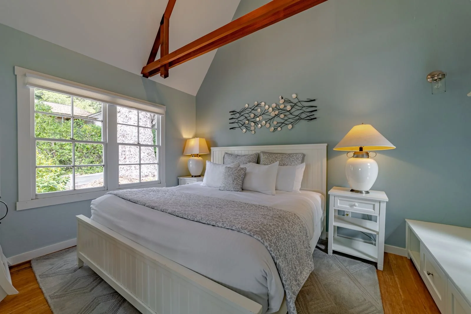 A cozy bedroom with light blue walls, a white bed with patterned and plain pillows, and matching nightstands with lamps. A large window with white frames lets in natural light, and a wooden beam decorates the ceiling. A metal wall art piece is mounte
