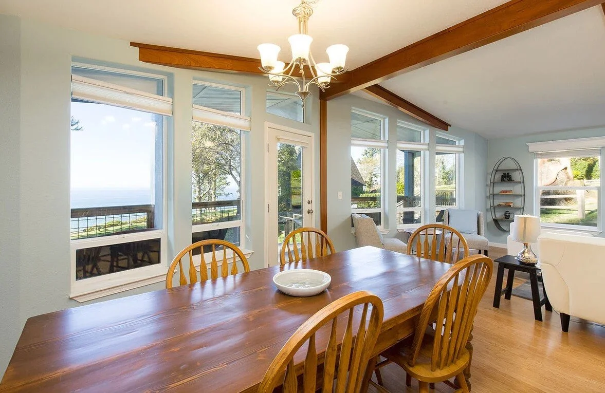 Bright dining and sitting area with large windows showing a scenic outdoor view, wooden dining table with chairs, chandelier, and cozy seating with sofa and armchair.