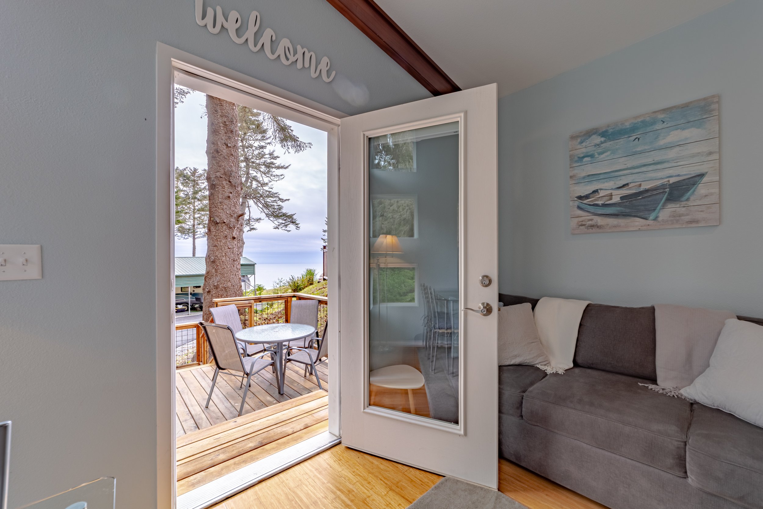 Living room with open door leading to a wooden patio overlooking a large tree and water, with a round table and chairs outside.