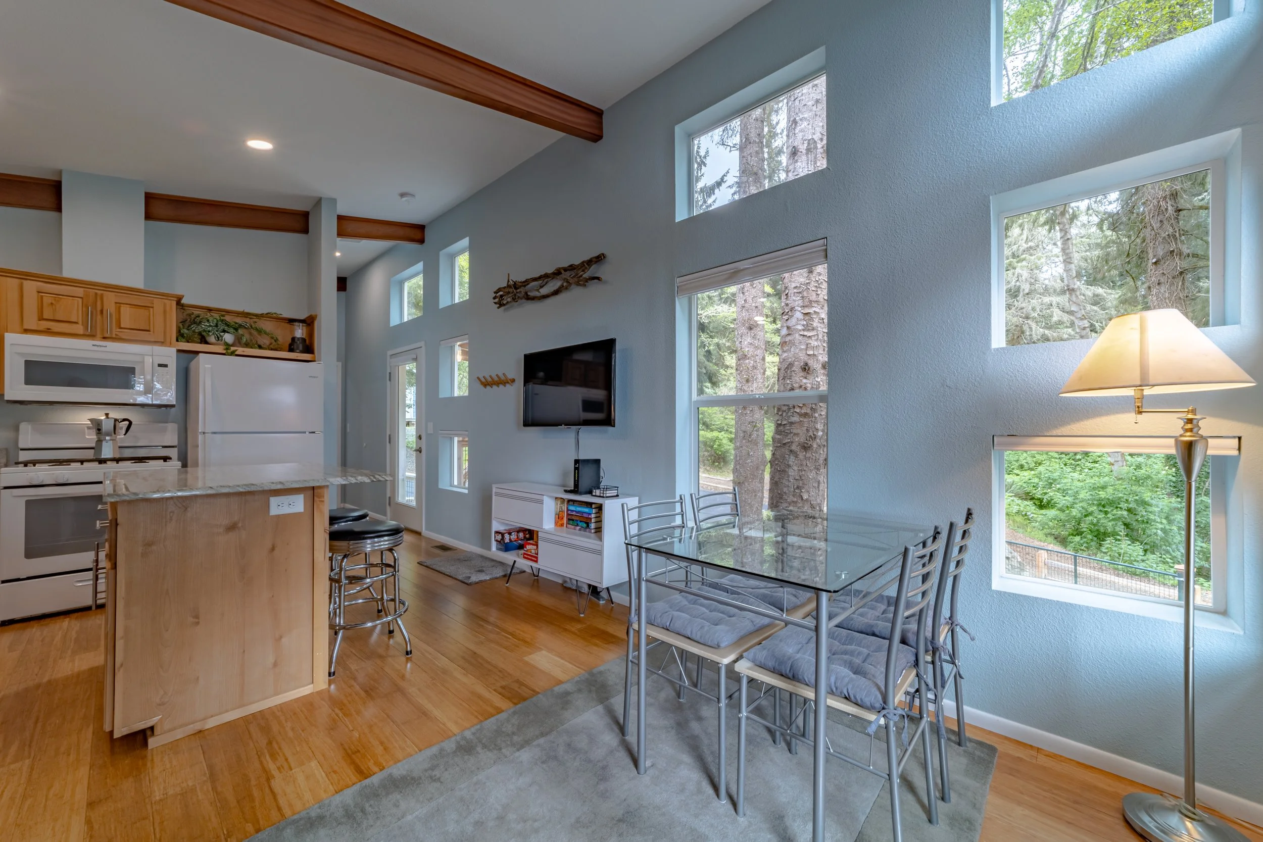 Interior view of a modern living and dining area with large windows showing trees outside. The space has light blue walls, wooden floors, a glass dining table with metal chairs, a TV mounted on the wall, and kitchen cabinetry and appliances visible o