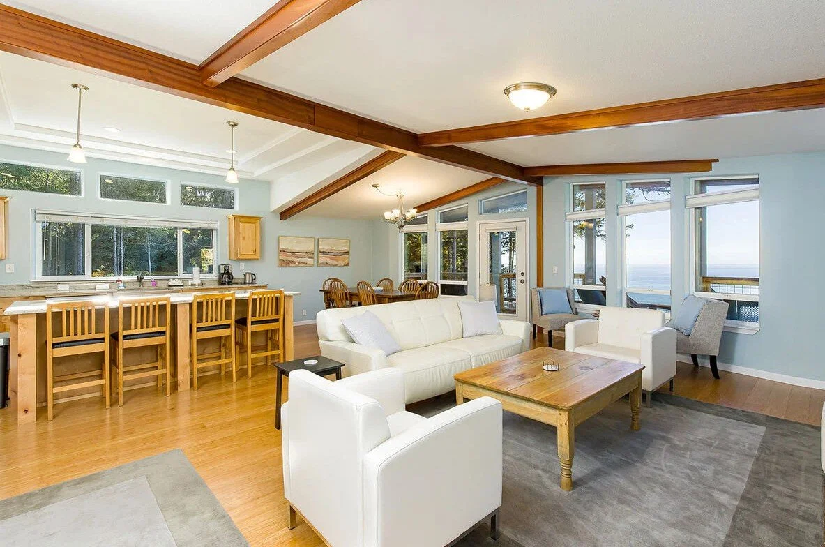 A bright and airy living room with large windows and a view outside. Contains white sofas, a wooden coffee table, a small side table, and a section of a kitchen with a breakfast bar and wooden chairs. Wood beams are on the ceiling, and there's a dini