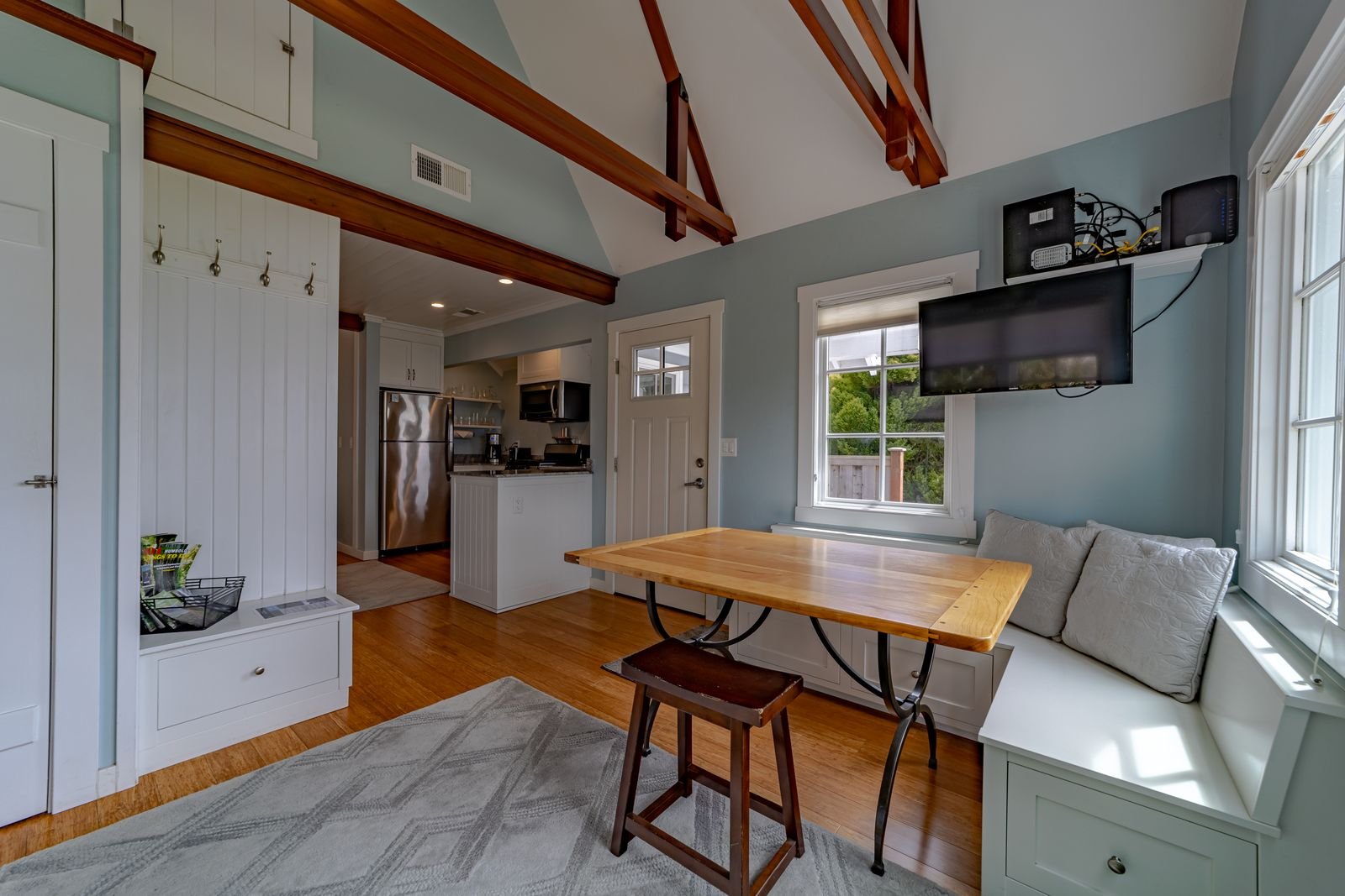 Interior of a cozy living space with a wooden dining table, window bench with pillows, wall-mounted TV, and view of the kitchen with stainless steel appliances.