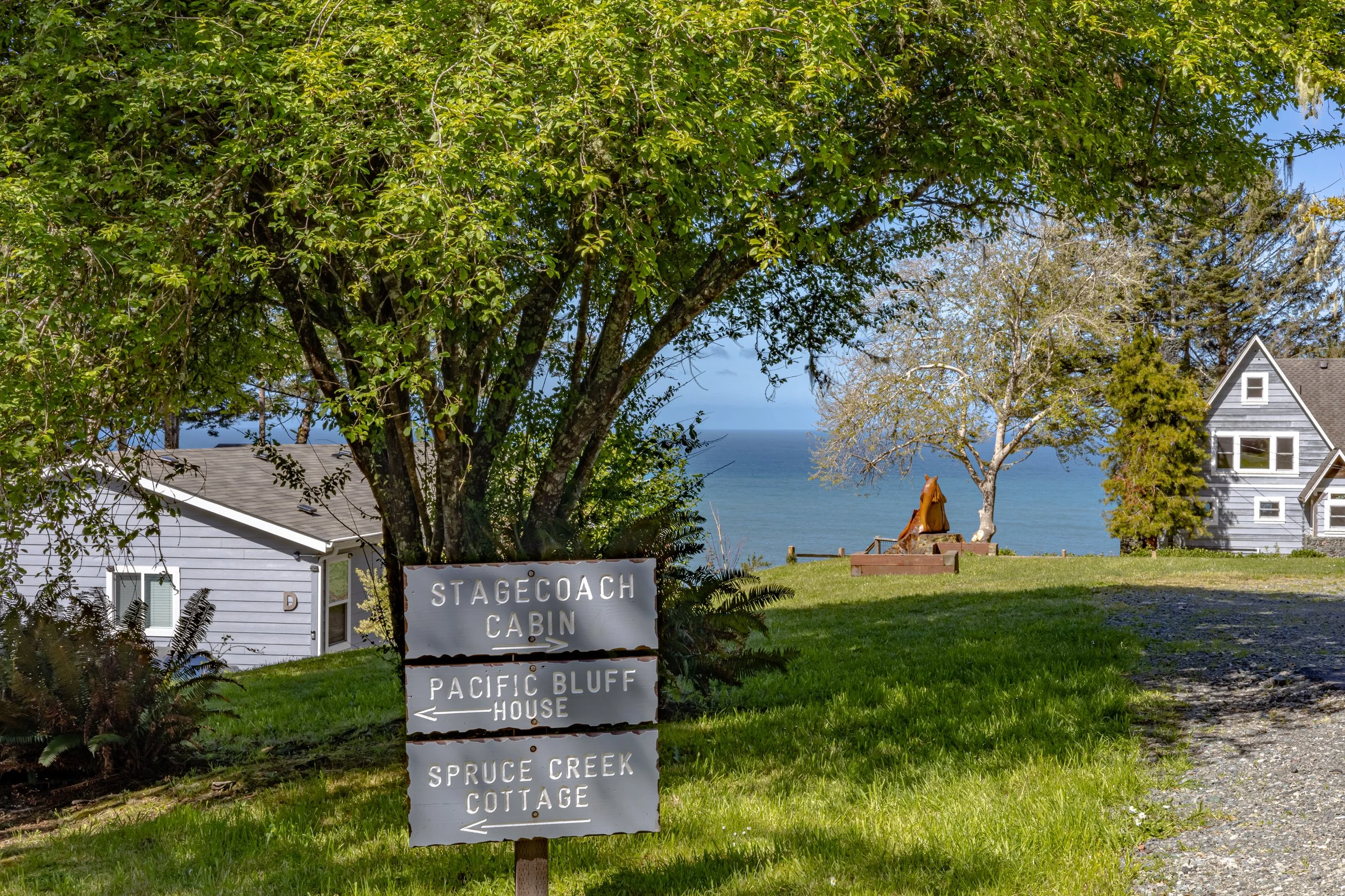 A sign in front of trees and houses points to the Stagecoach Cabin, Pacific Bluff House, and Spruce Creek Cottage, with a view of the ocean in the background.