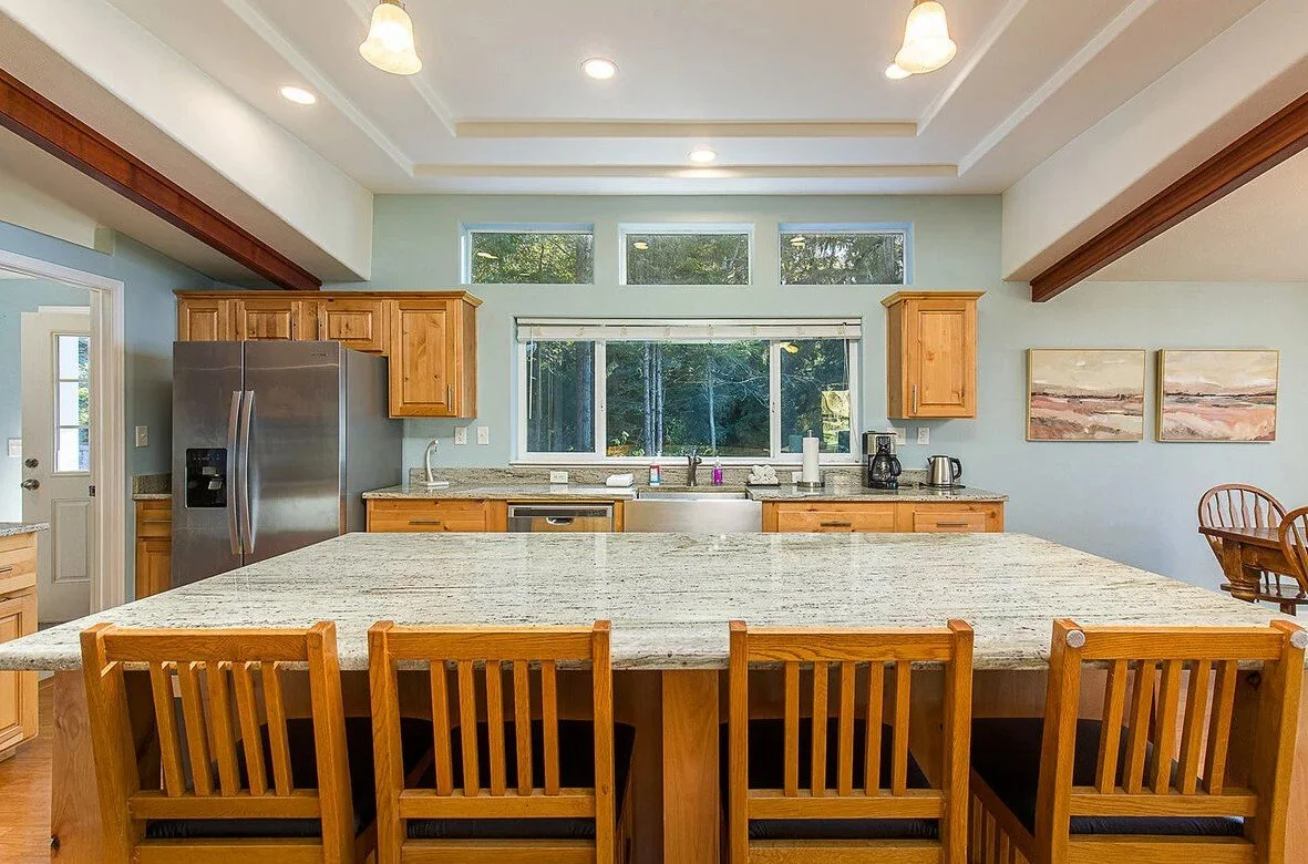 Kitchen with a large granite countertop island, wooden cabinets, stainless steel refrigerator, and a window above the sink.