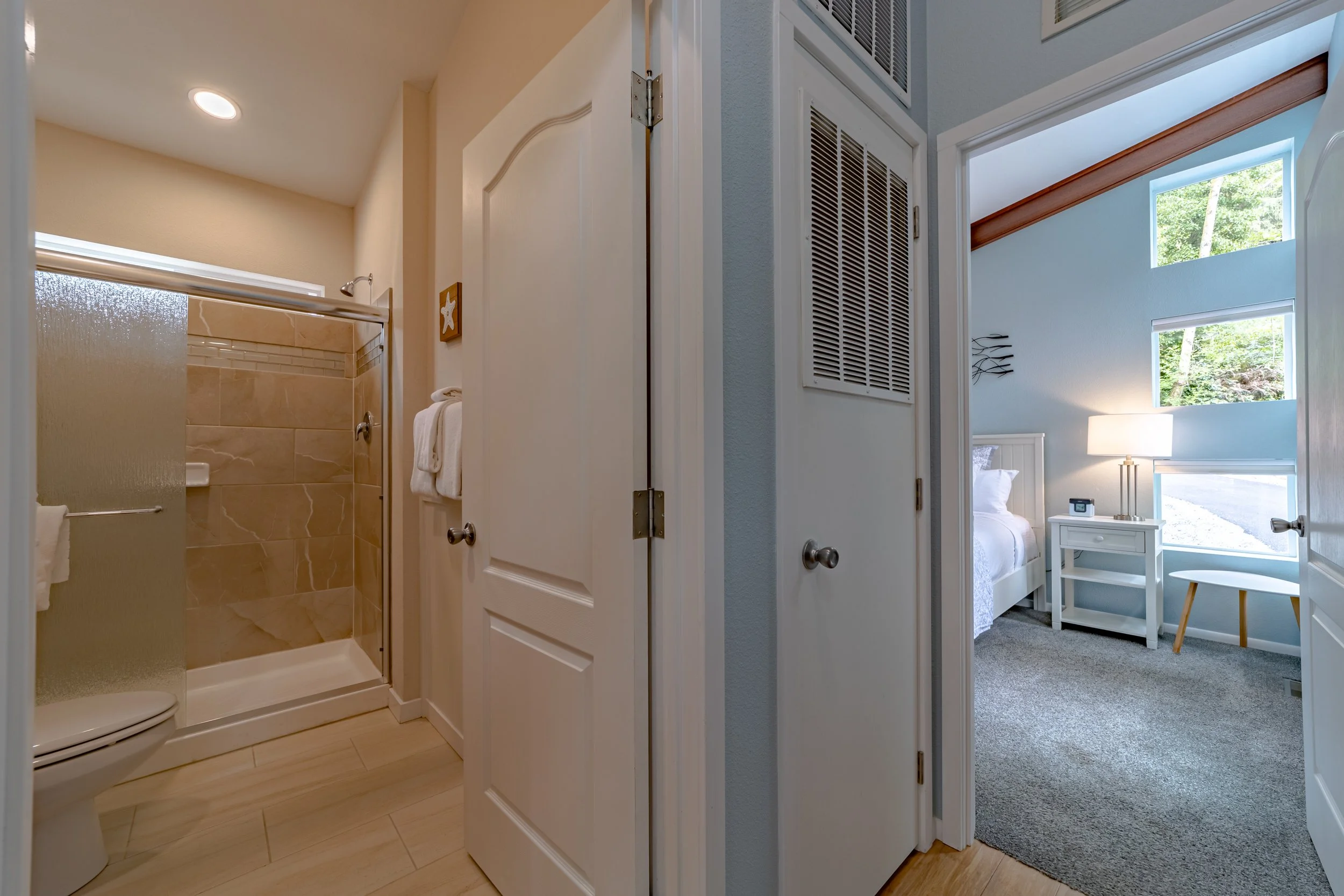 View of a bathroom with a shower stall, towels hanging, and beige tiled walls; adjacent bedroom with white bed, nightstand, lamp, and large windows showing outdoor greenery.