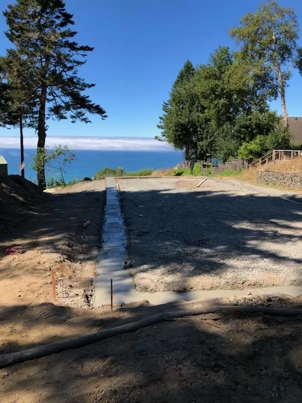 Construction site with a freshly poured concrete slab on a dirt surface, overlooking a body of water with a clear blue sky and trees nearby.