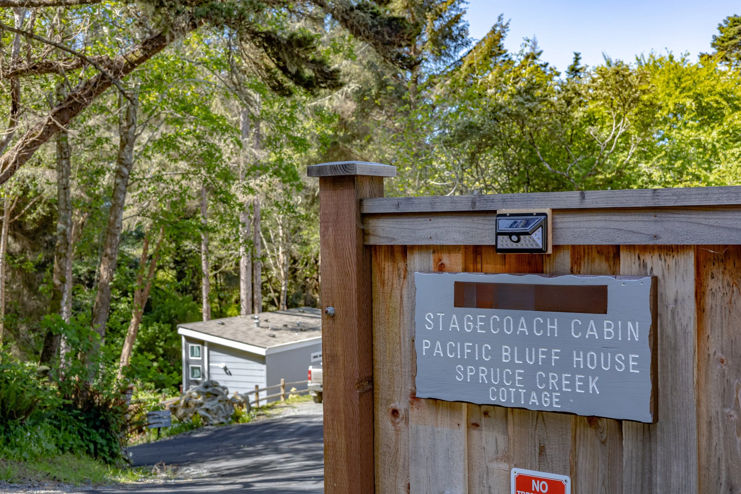 Close-up of a wooden entrance gate for a cabin, with a sign listing 'Stagecoach Cabin, Pacific Bluff House, Spruce Creek, Cottage,' and a small security camera mounted above the sign. The background shows green trees and a small building with a drive