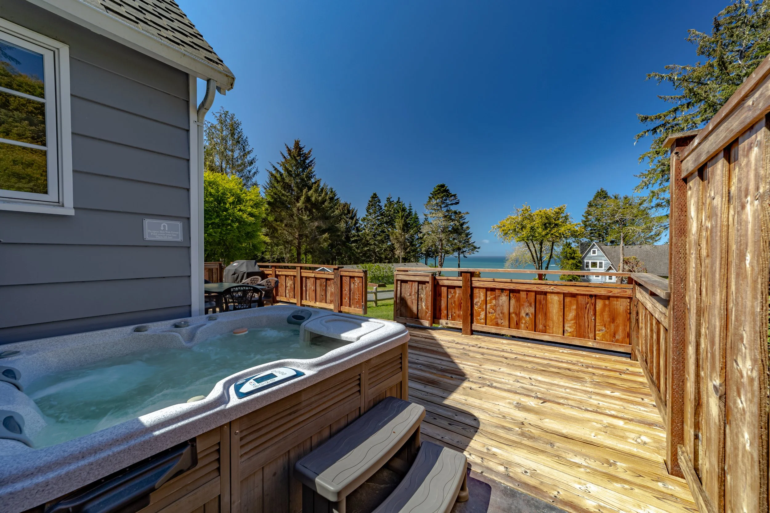 Outdoor patio with hot tub, wooden deck, and view of trees and ocean in the distance under a bright blue sky.