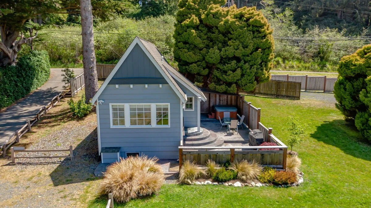 A small blue house with a gabled roof, surrounded by lush green trees and a fenced yard with outdoor furniture and a hot tub.