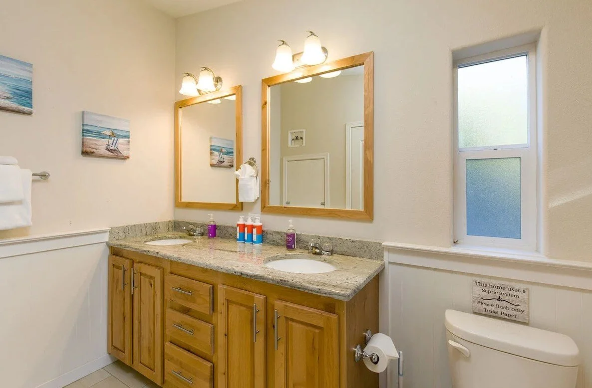 A bathroom with a double wooden vanity, granite countertop, and two mirrors. There are three bottles of soap or lotion on the counter. Above the mirrors are two light fixtures with three bulbs each. A small window is on the right, and a toilet is vis