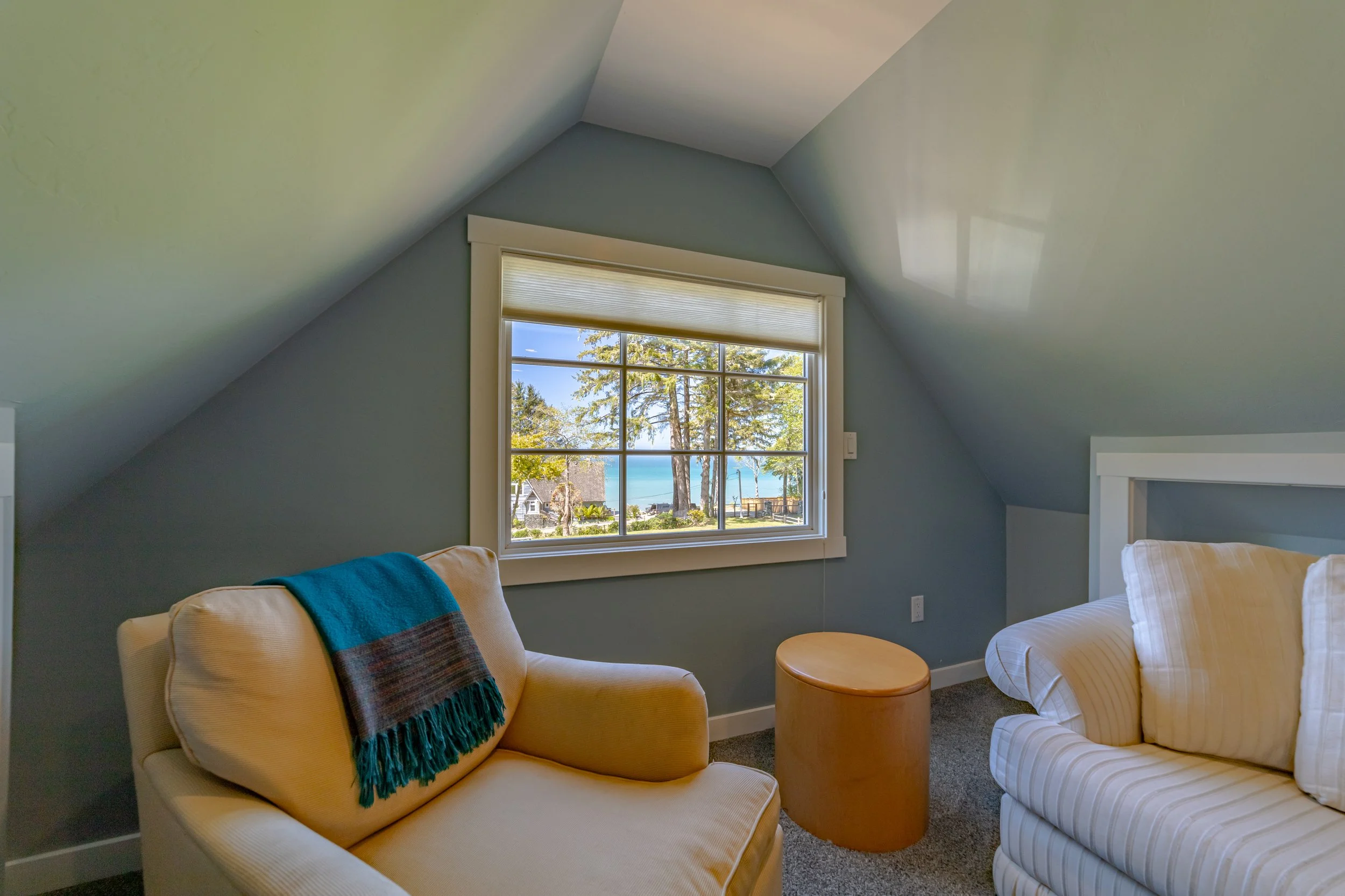 A cozy attic room with a large window showing a view of trees and the ocean. The room has a beige armchair with a blue and brown throw blanket, a striped white sofa, a small wooden side table, and soft natural lighting.