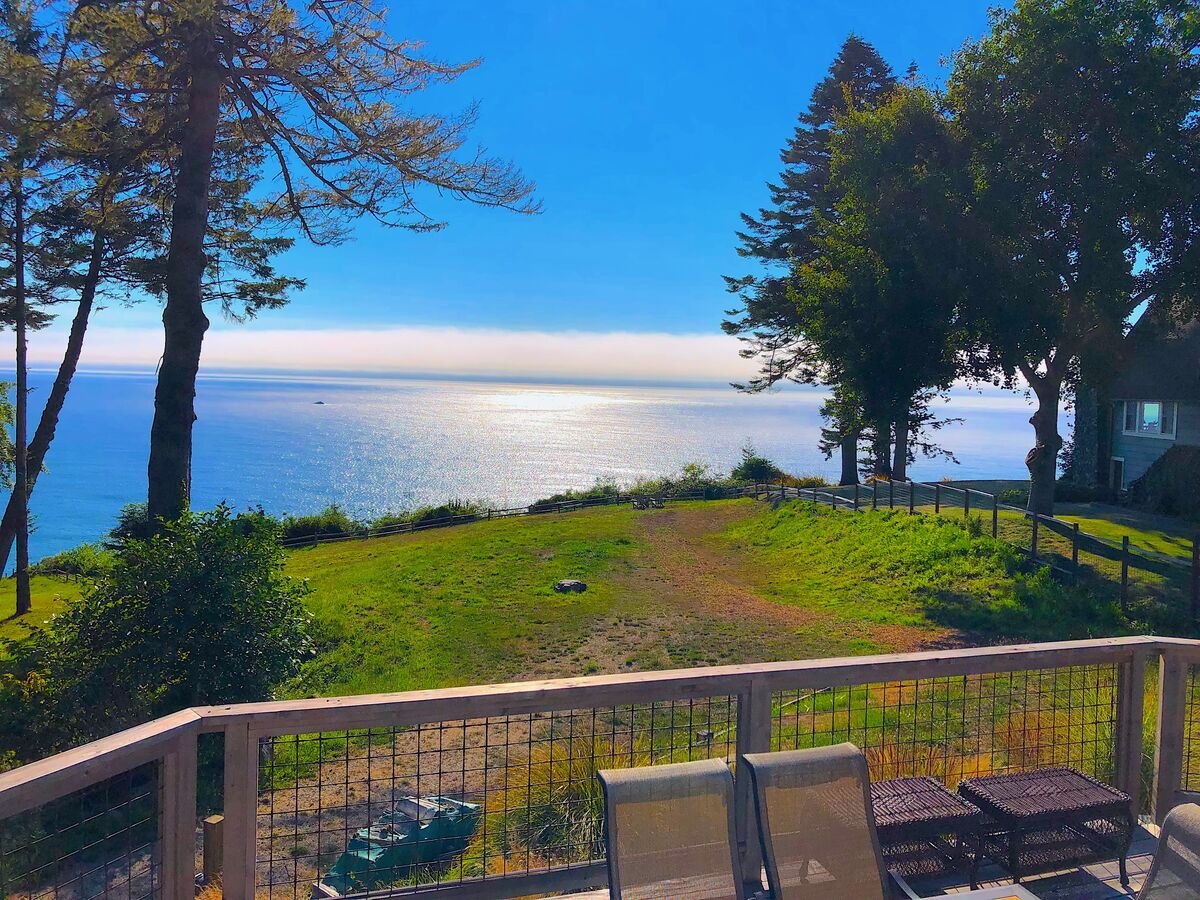 Ocean view from a balcony with patio furniture, trees, and a grassy area, under a bright blue sky.