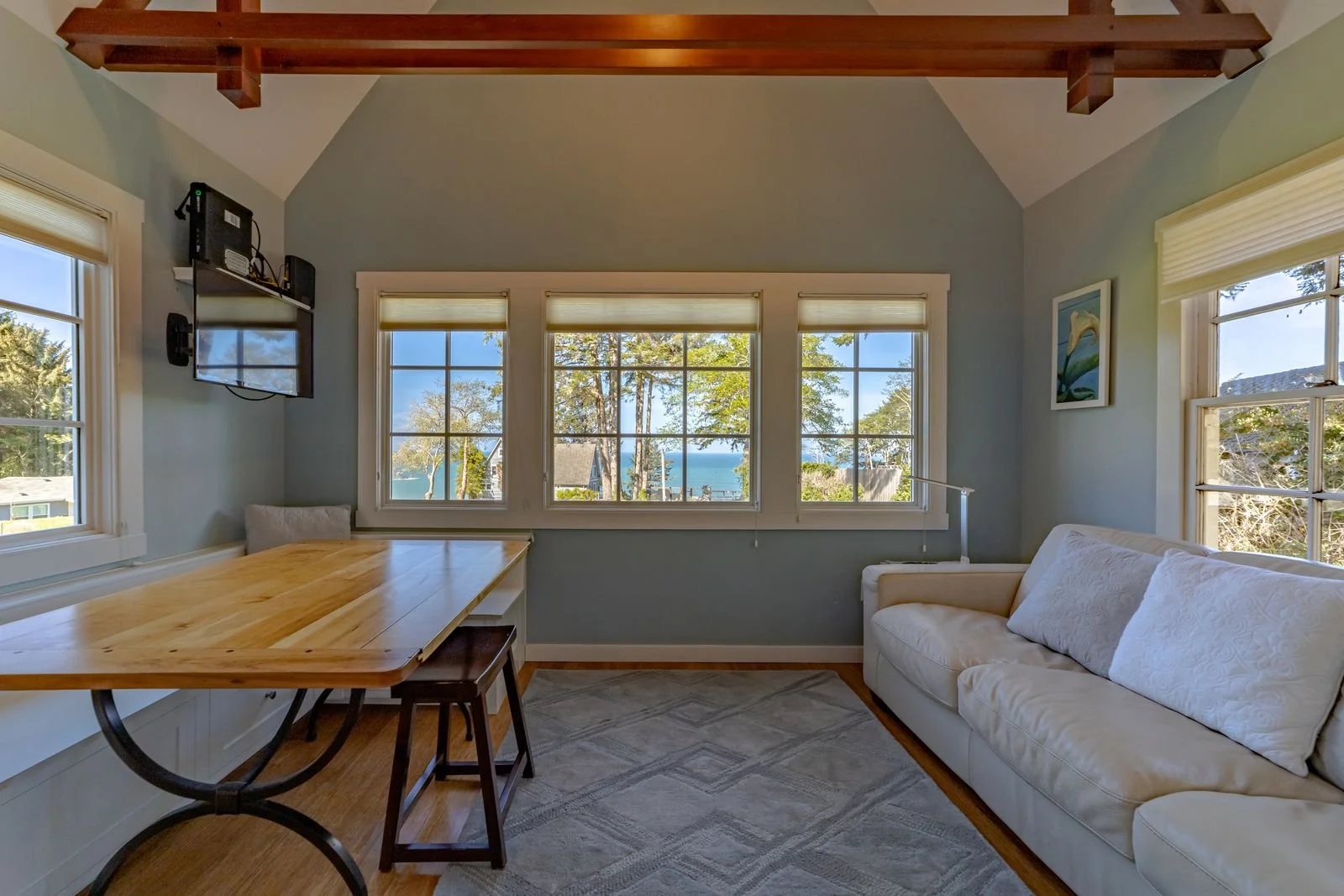 Living room with three large windows showing trees and a view of the water, a white couch with four pillows, a wooden table, a TV mounted on the wall, and a decorative wall art.