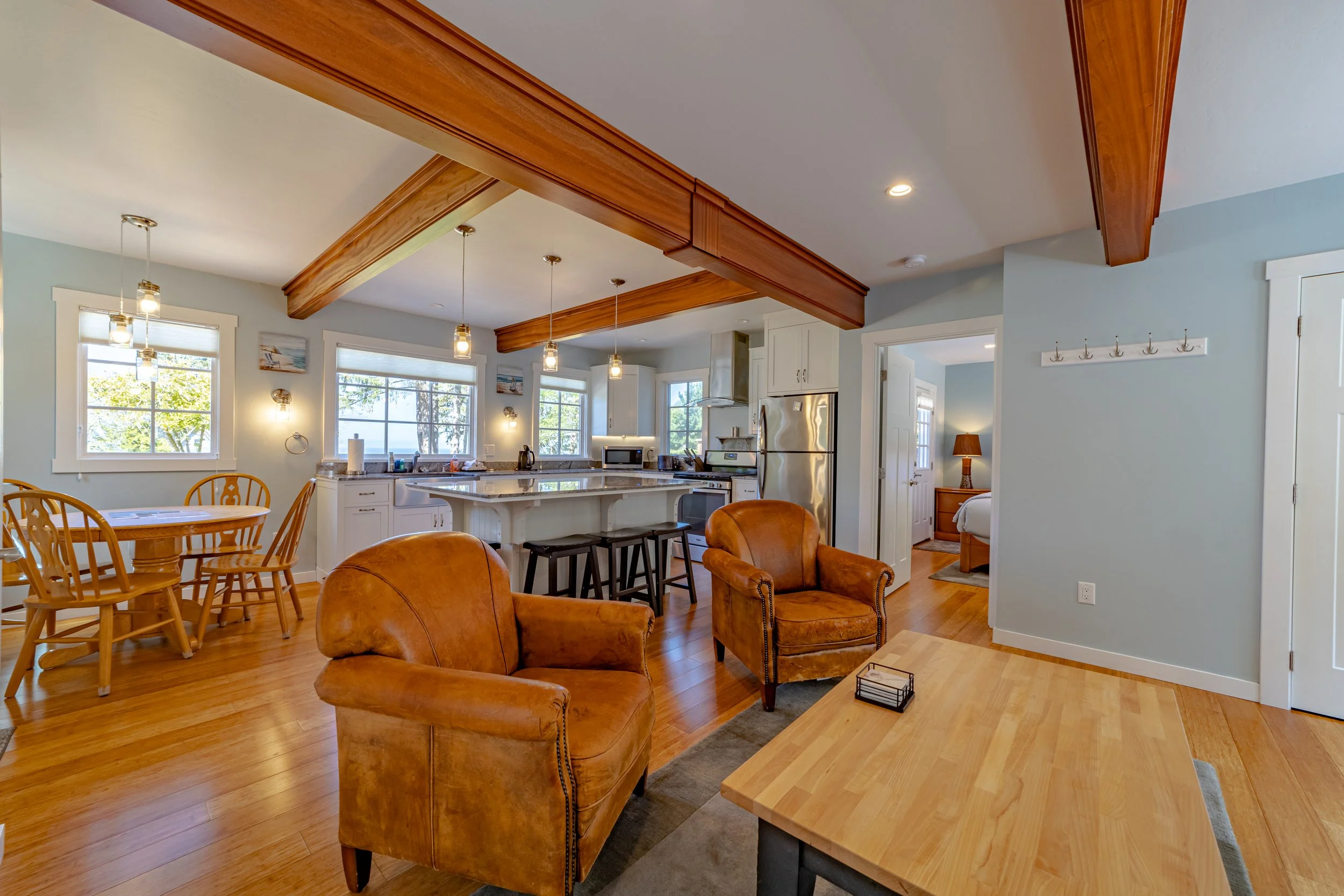 Open-concept living room and kitchen with hardwood floors, leather armchairs, wooden dining table, ceiling beams, pendant lights, and large windows with a view of trees outside.