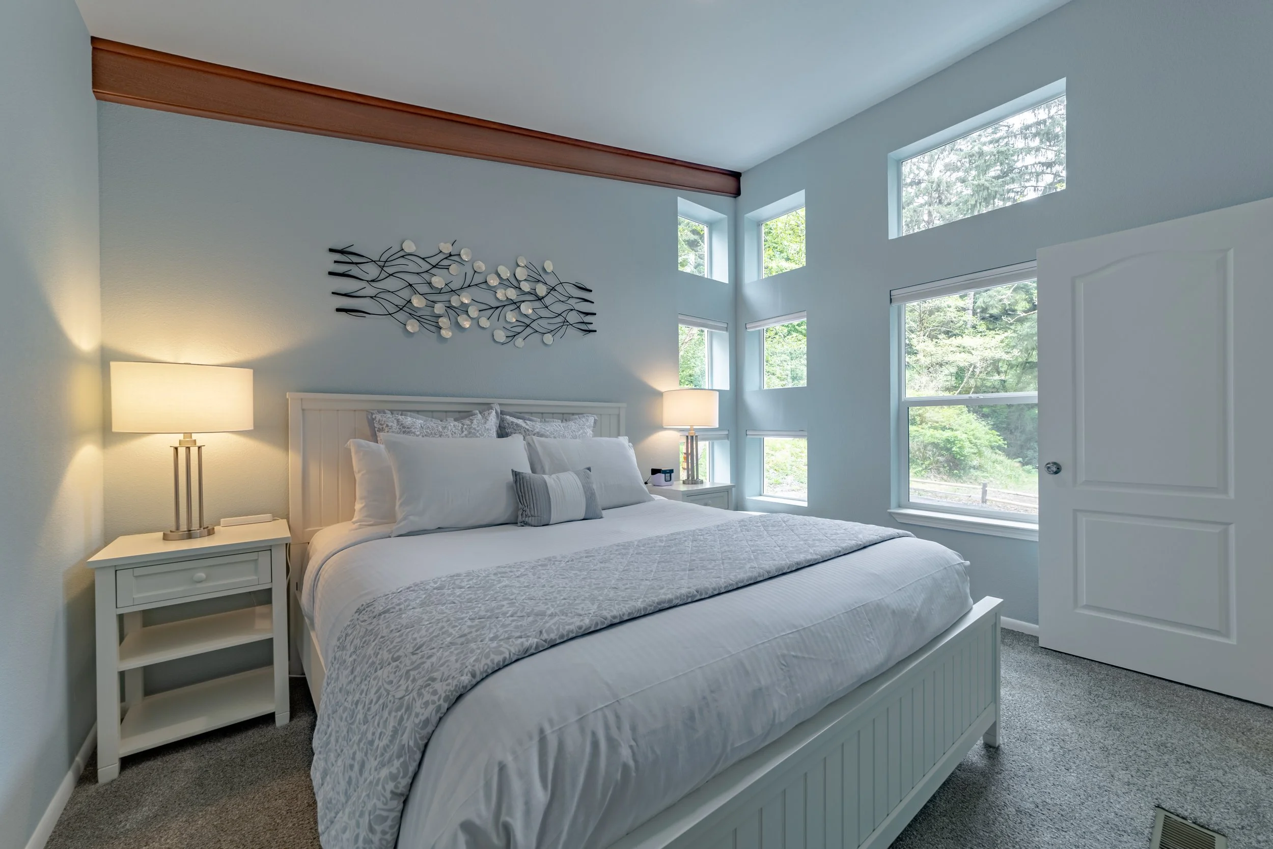 Bedroom with white bed, two bedside tables with lamps, decorative wall art above bed, large windows showing greenery outside.