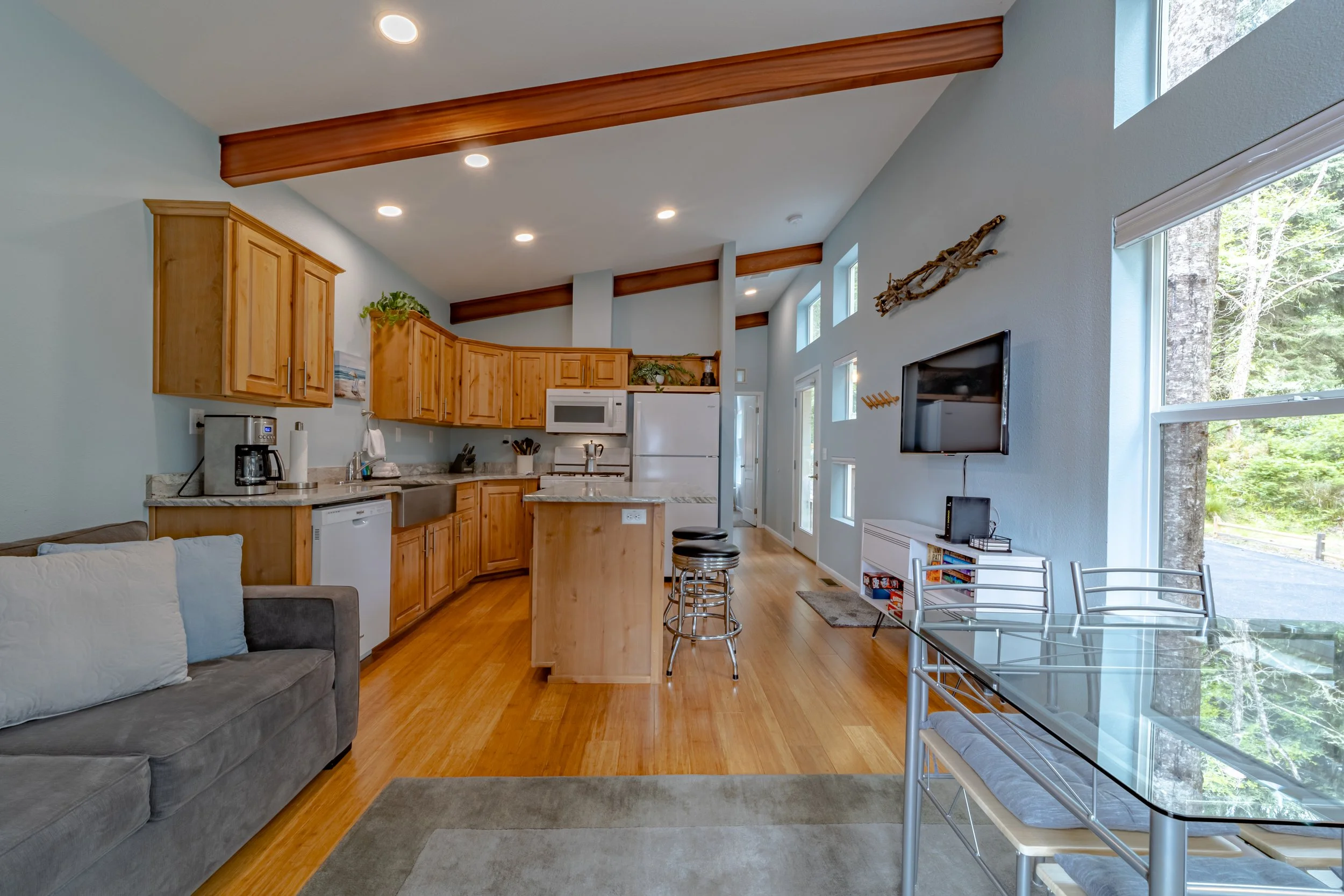 Open-concept living room and kitchen with wooden cabinetry, light blue walls, hardwood floors, and large windows showing trees outside.