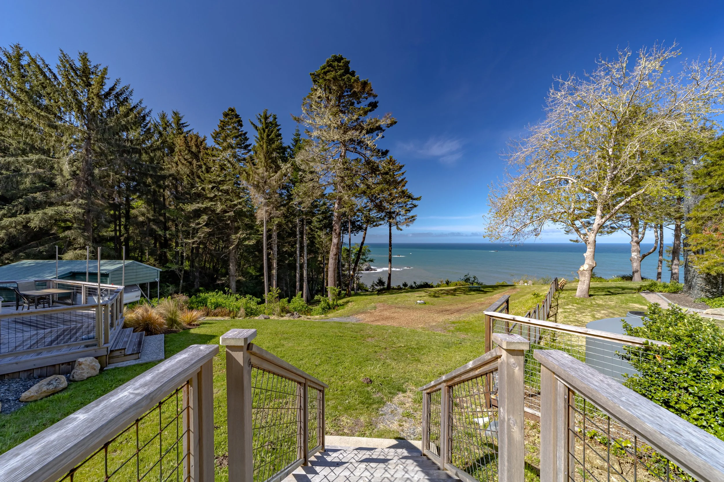 View from a wooden staircase leading to a grassy yard with trees, overlooking the ocean with blue sky and clouds.