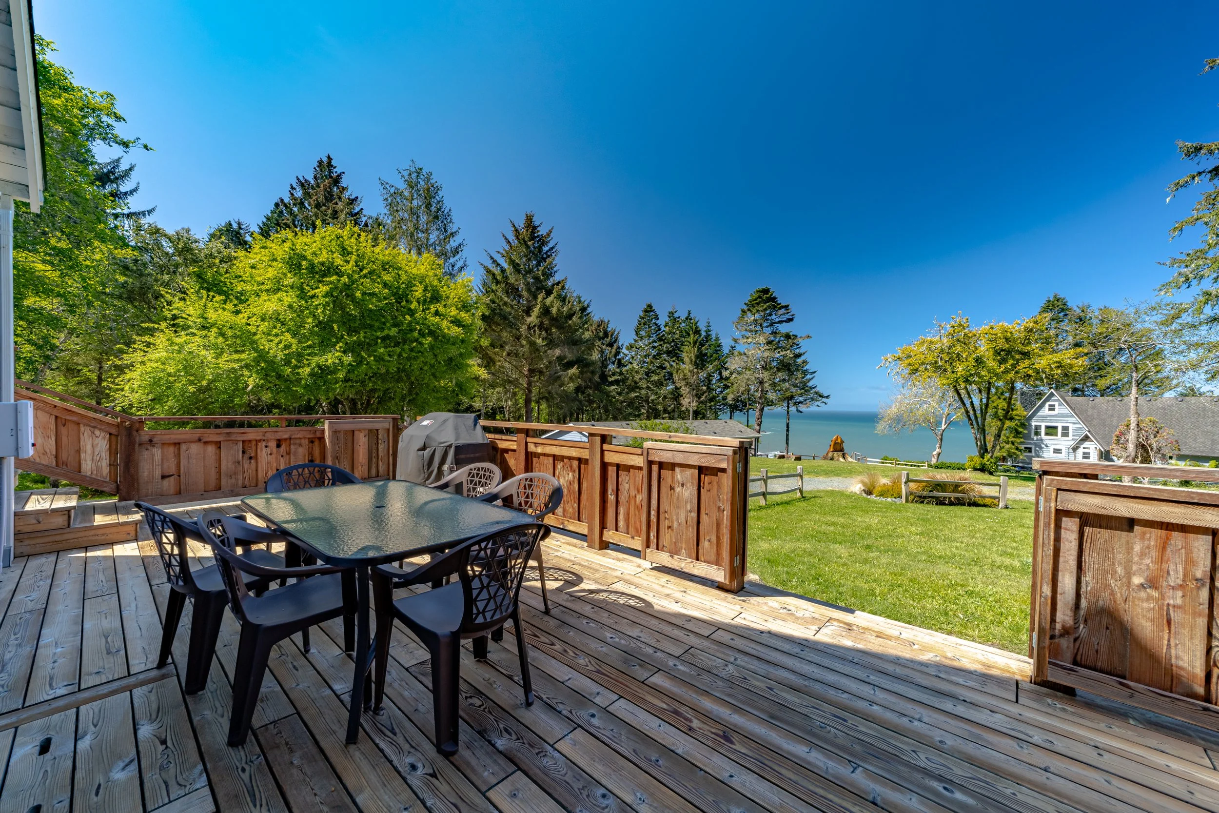 Wooden deck with black outdoor table and chairs, barbecue grill, overlooking green lawn, trees, and a distant view of the ocean and houses under a clear blue sky.