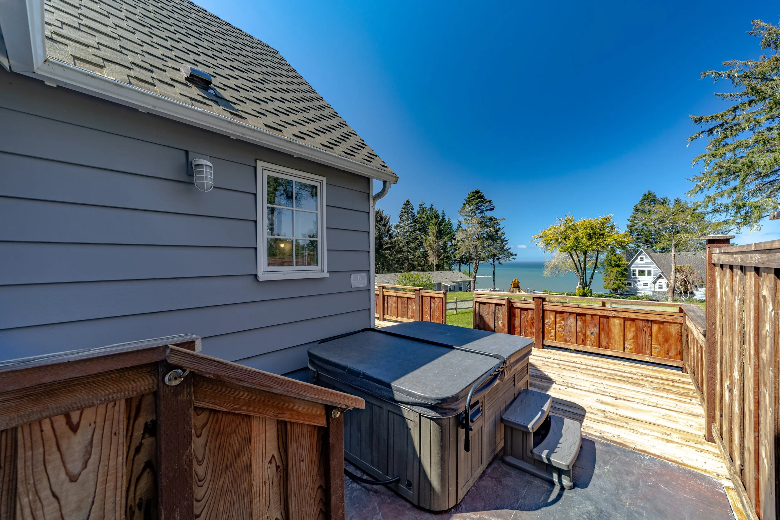 View of a backyard deck with a hot tub and a scenic ocean view, various trees, and neighboring houses under a bright blue sky.
