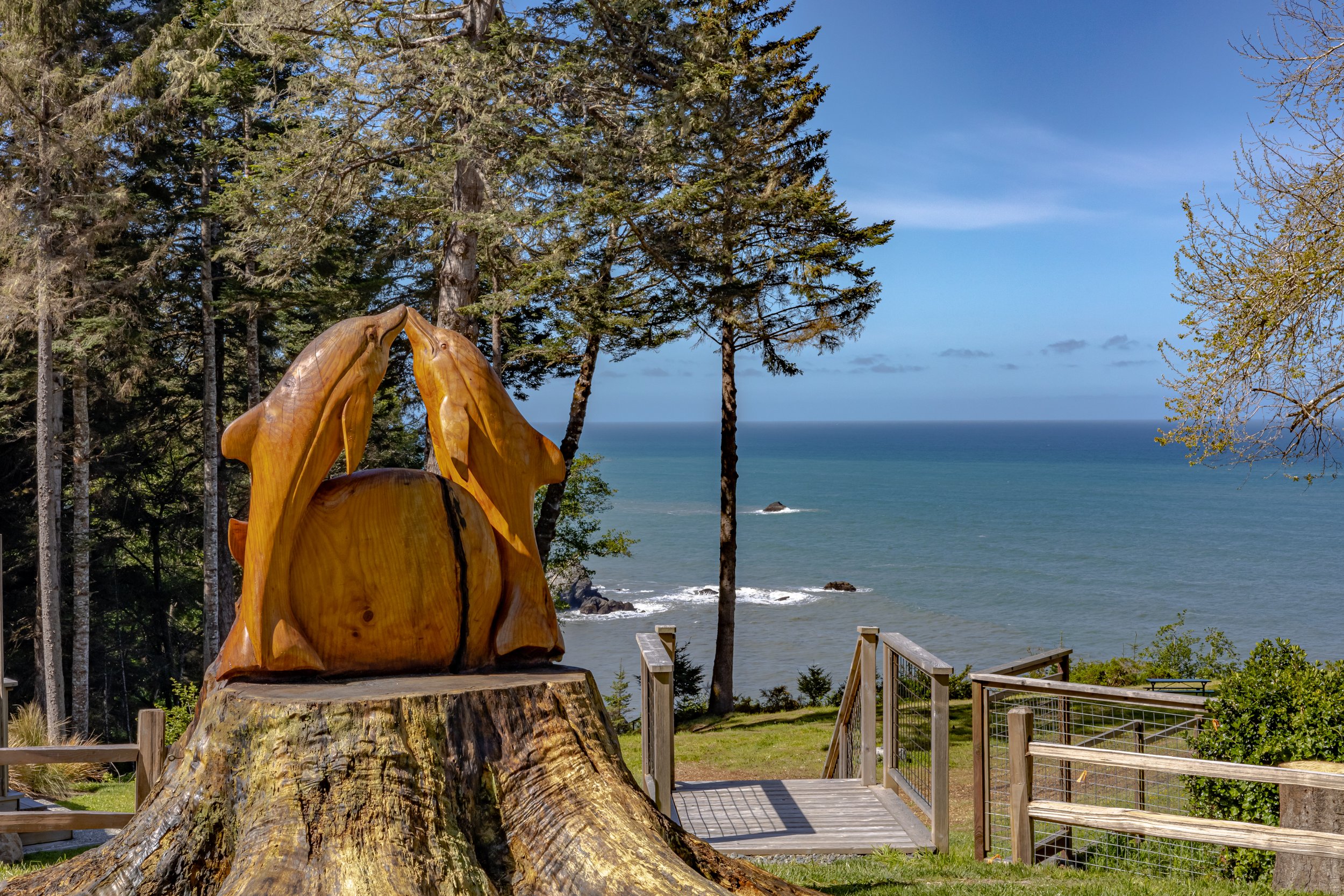Wood sculpture of two horses nuzzling, mounted on a tree stump, near a pathway with a railing, overlooking the ocean with trees and a blue sky in the background.