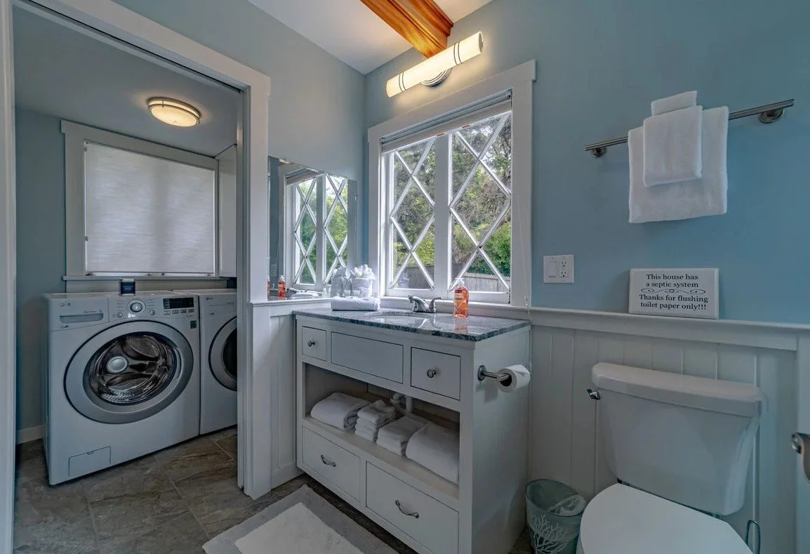 Bright laundry room with washing machine and dryer, a white vanity with a sink, a window with diamond-shaped panes, a towel bar with a towel, and a toilet. There is a sign indicating the house has a septic system. The room has a light blue wall with 