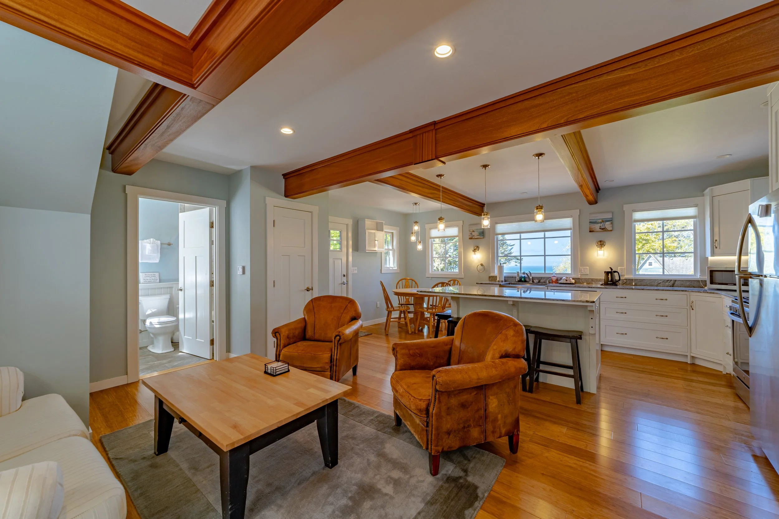 Living room and kitchen area with hardwood floors, light-colored walls, wooden ceiling beams, and modern lighting fixtures. The living room has a beige sofa, two brown armchairs, and a wooden coffee table. The kitchen features white cabinets, a marbl