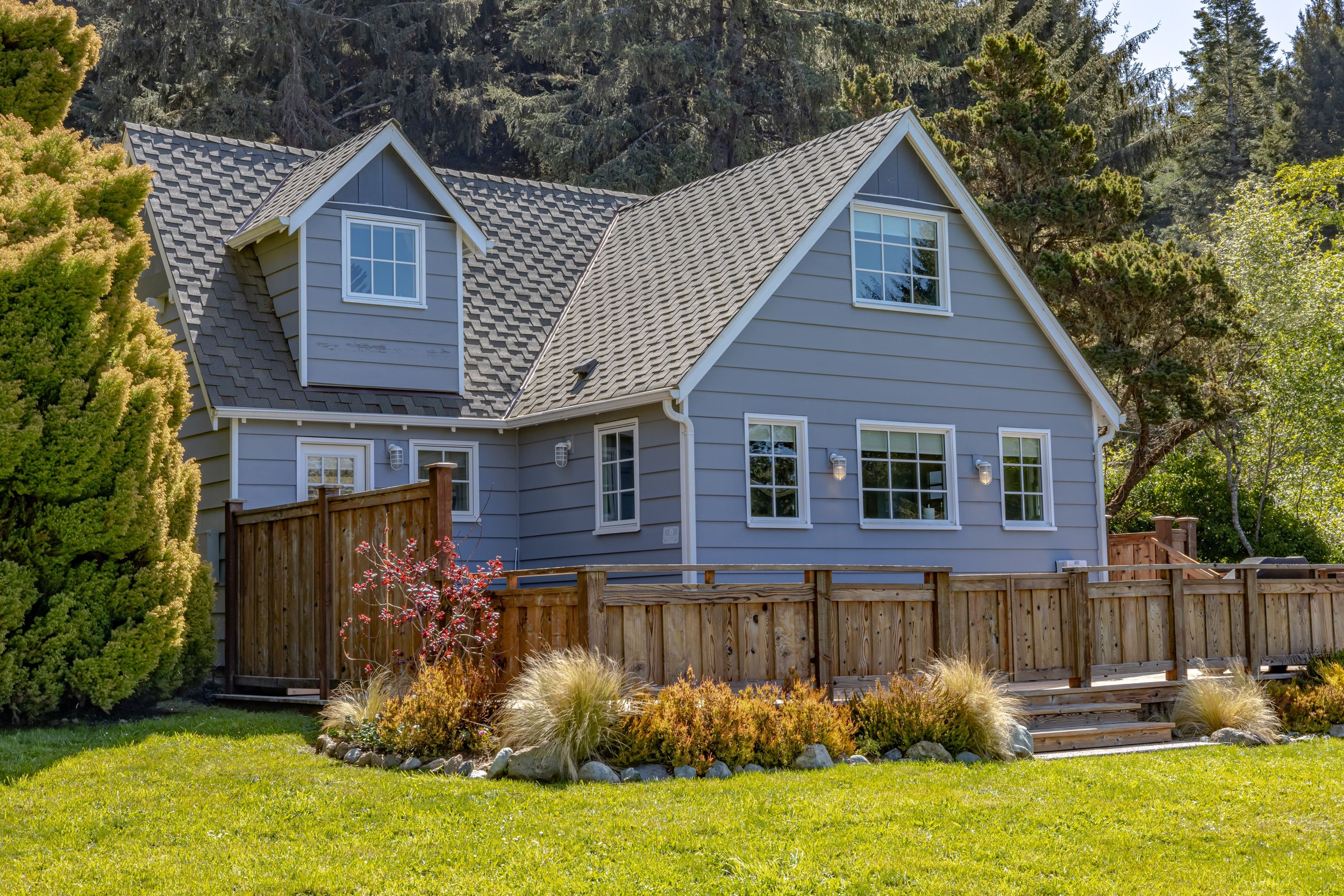 A two-story house painted in light blue with white trim, featuring multiple windows and a gray tiled roof, surrounded by green trees and a wooden deck in a landscaped yard.