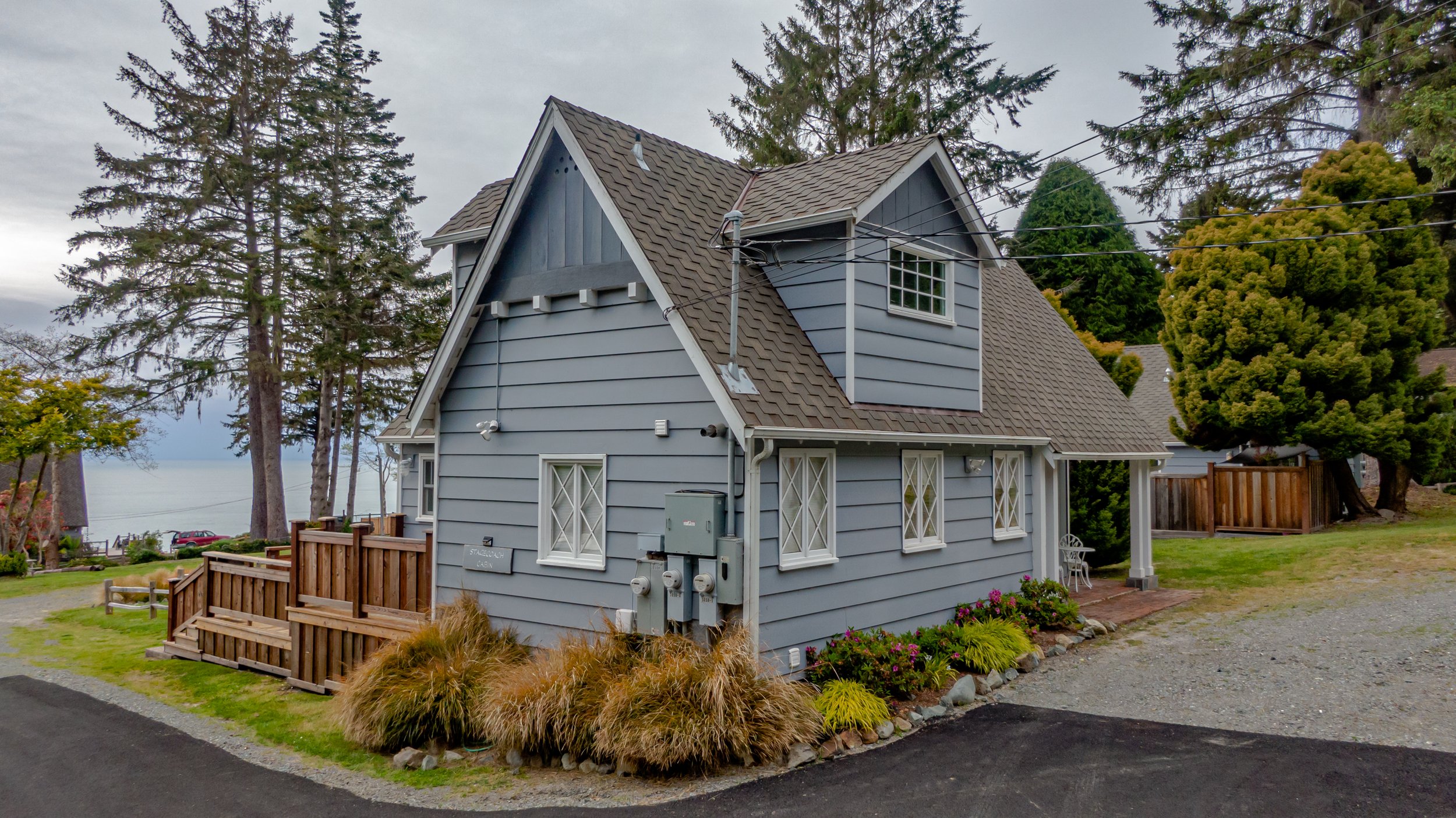 A two-story blue house with brown shingle roof, white trim, and multiple windows, situated near water with tall trees and a grassy area with plants and flowers.