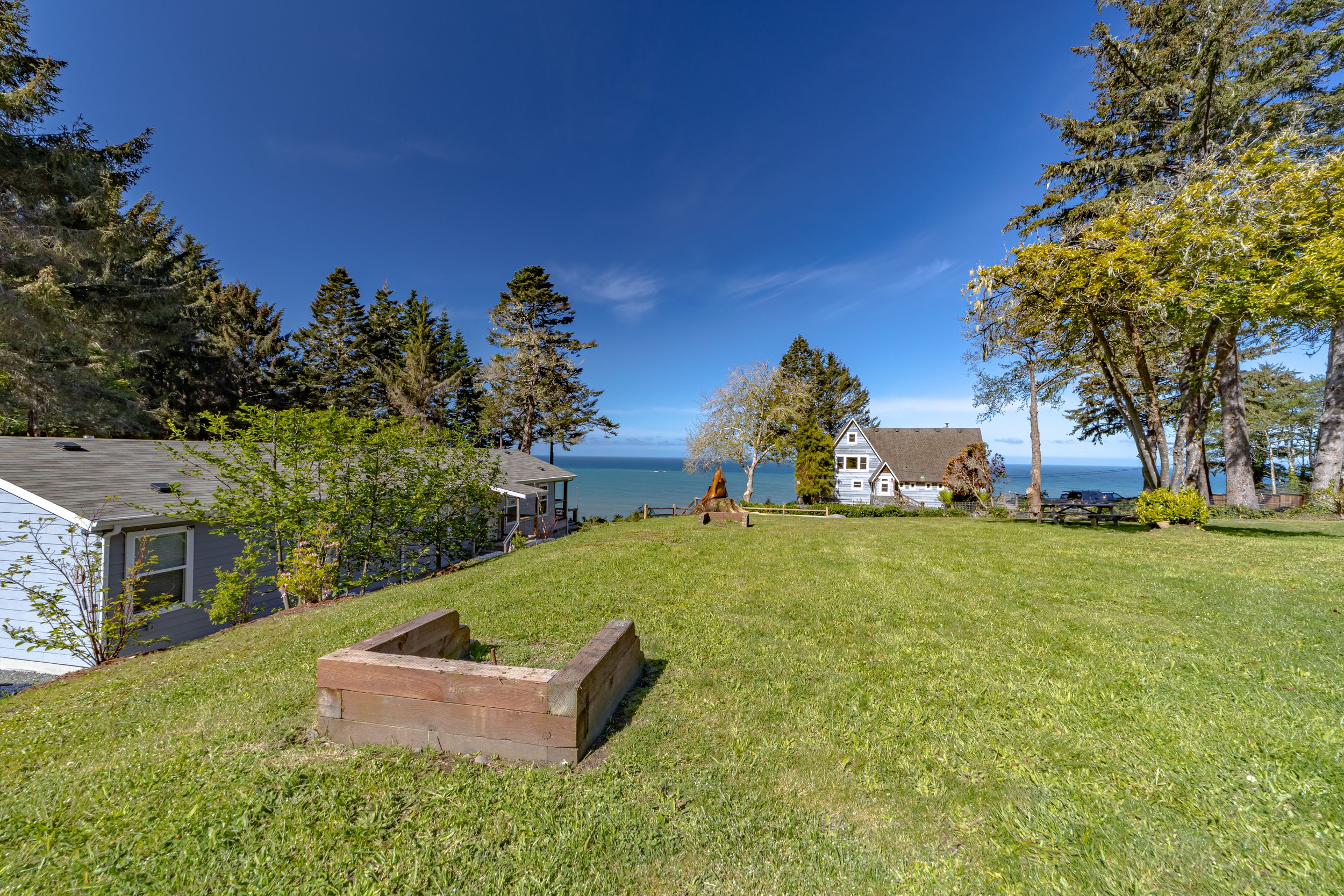 A scenic view of a green grassy yard with trees, a house, and the ocean in the background under a blue sky.