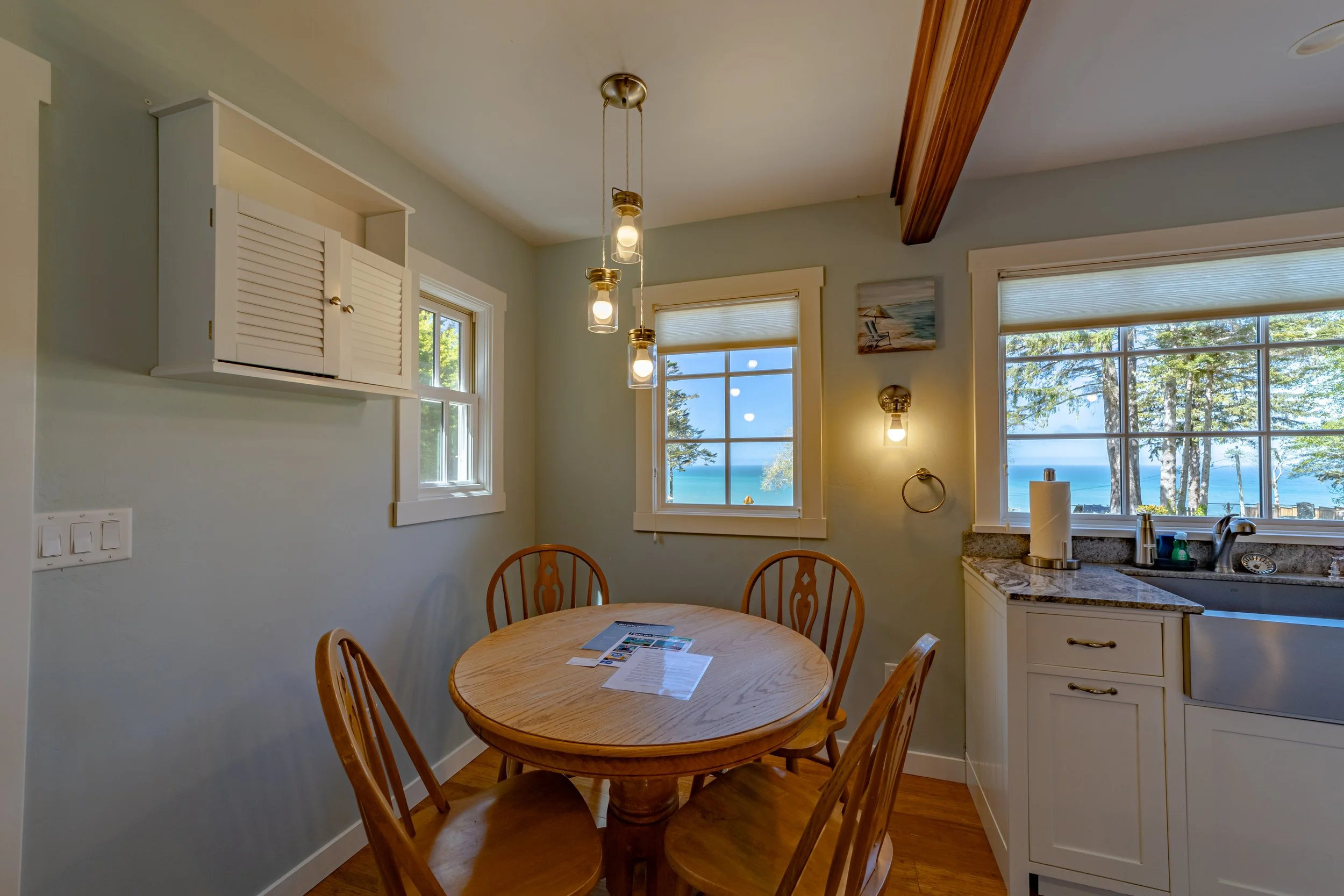 Dining area with a round wooden table and four wooden chairs, three windows with views of trees and water, and a kitchen counter with a paper towel roll, soap, and dishes, in a cozy home.