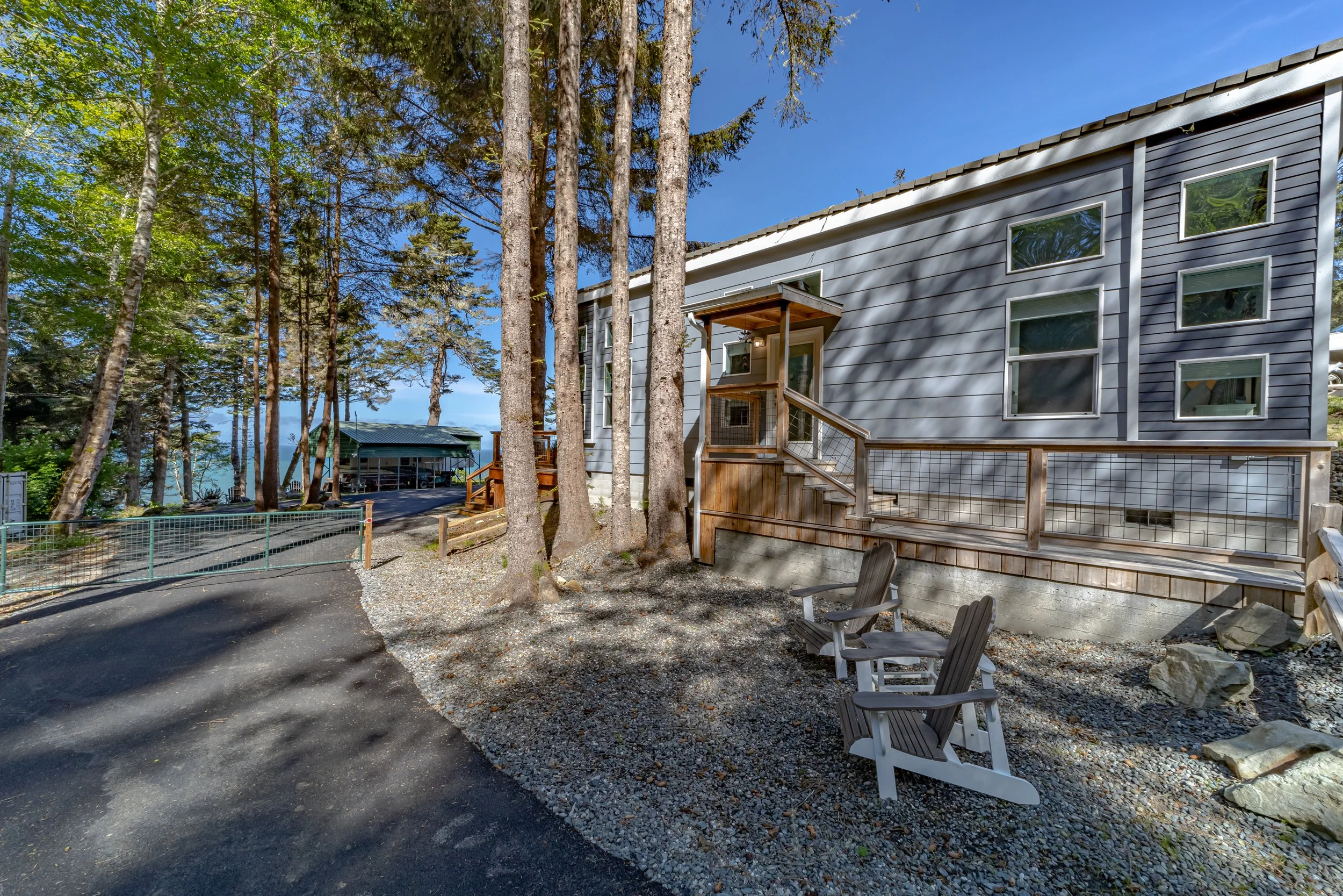 A house with grey siding and multiple windows, surrounded by tall trees. There are two Adirondack chairs on a gravel patch in front of the house. A small wooden staircase leads to a back door, and a driveway runs nearby, with a small structure and a 