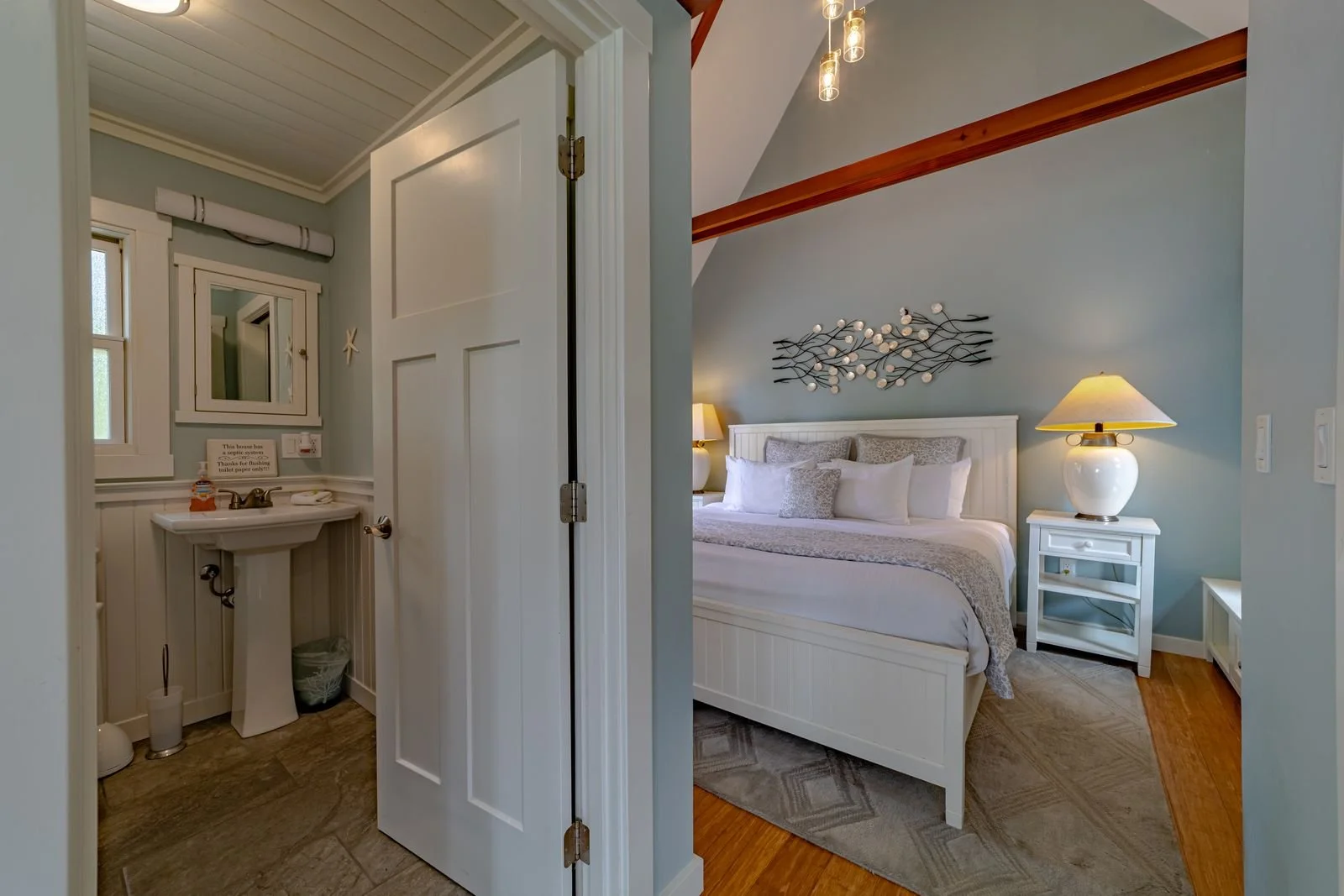 View of a bedroom with a white bed, nightstand, lamp, and wall art, and an adjoining bathroom with a small sink, mirror, and window.