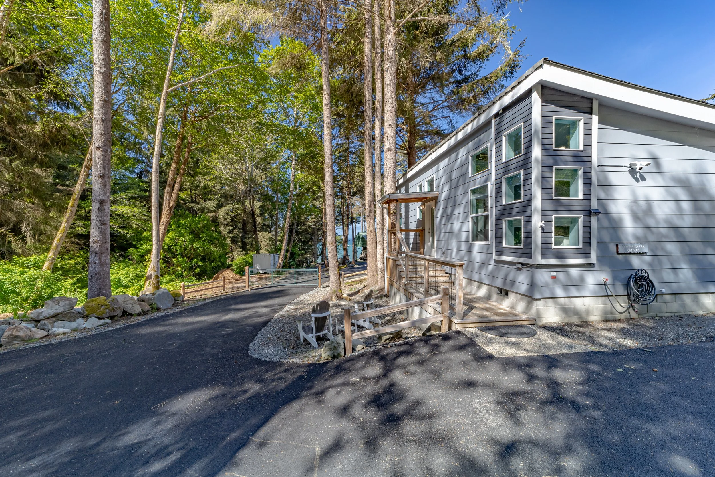 A modern house with multiple small windows situated on a wooded lot, with a paved driveway and two wooden chairs near the entrance. Tall trees surround the property, providing shade, and a rock-bordered garden area is visible on the left.