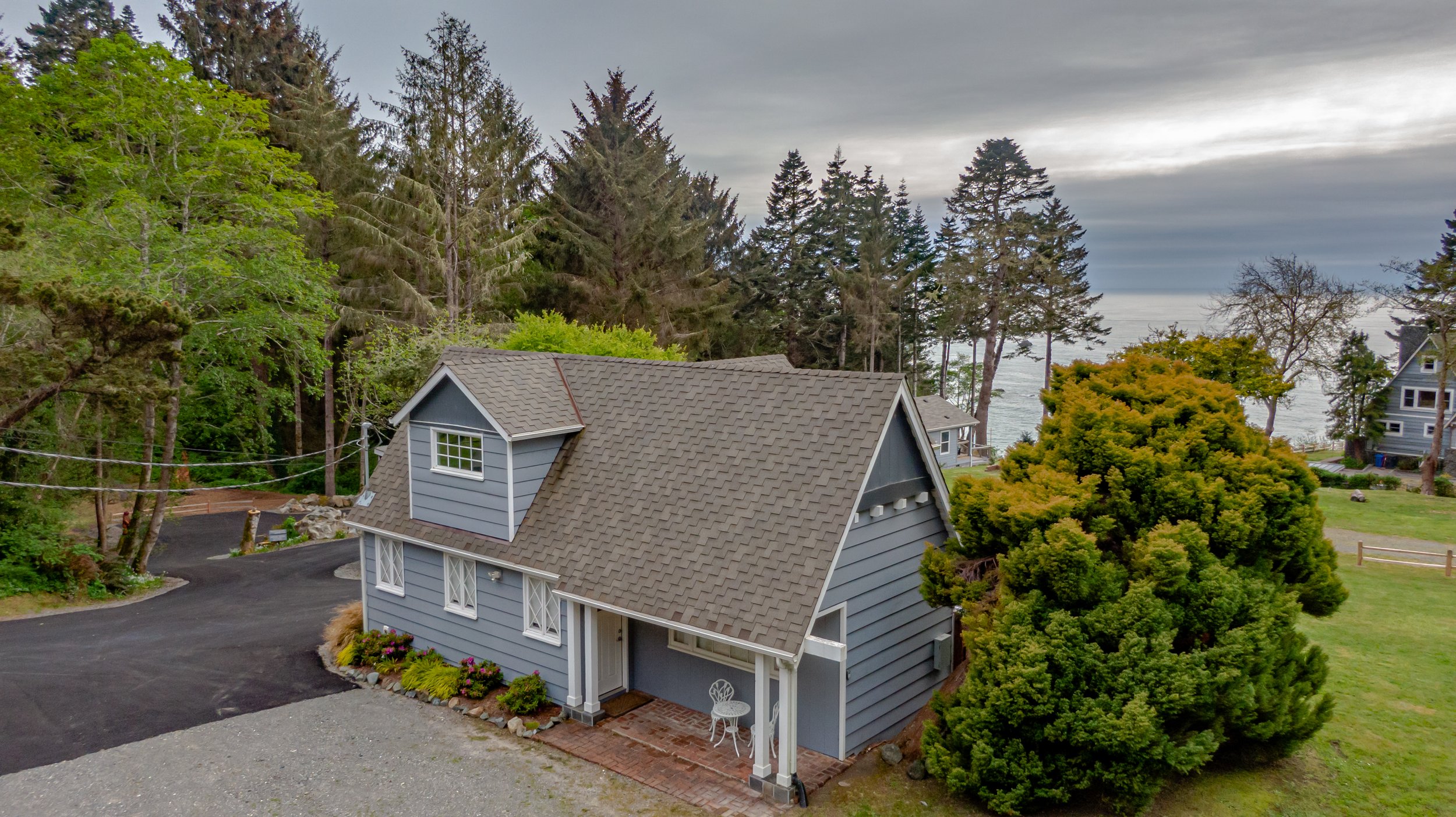 A blue house with a gabled roof and dormer window, surrounded by trees and greenery, with a view of the ocean in the background.