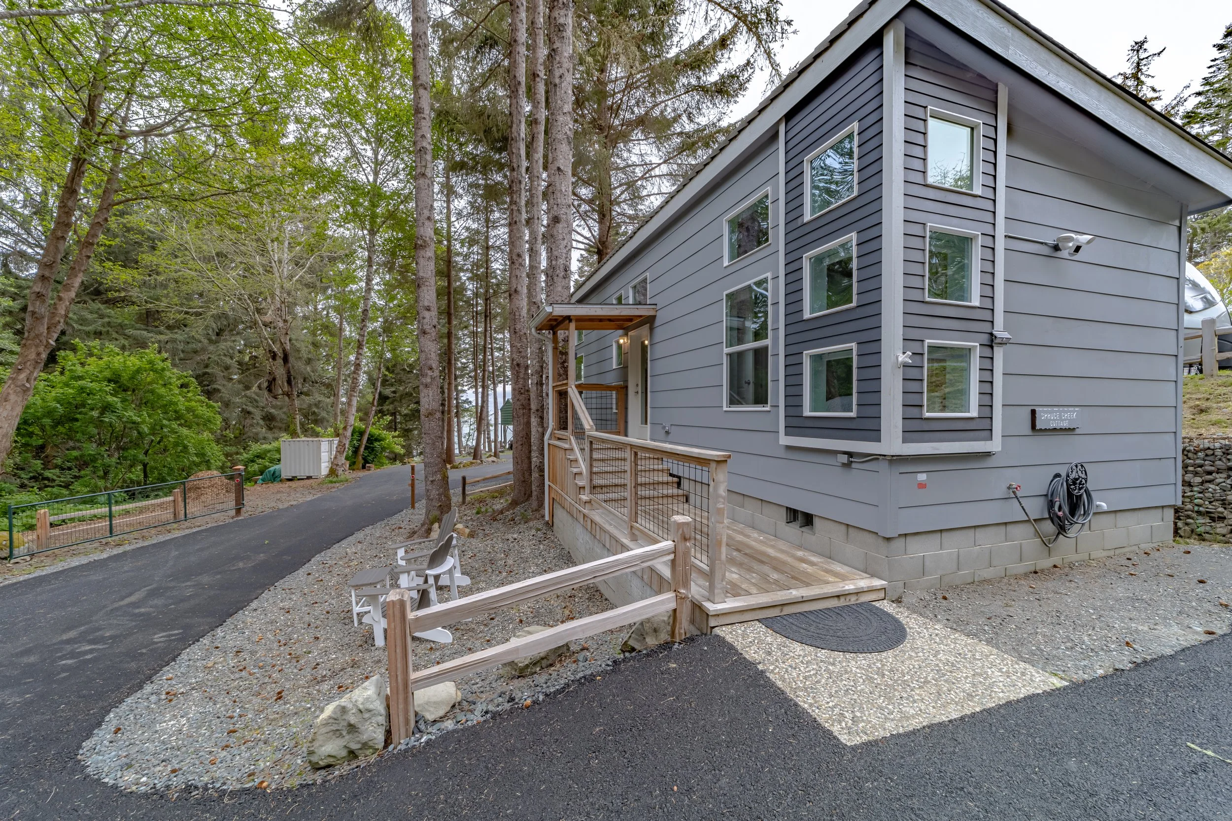 A modern gray house with multiple windows in a wooded area, featuring a small wooden ramp and chairs outside.
