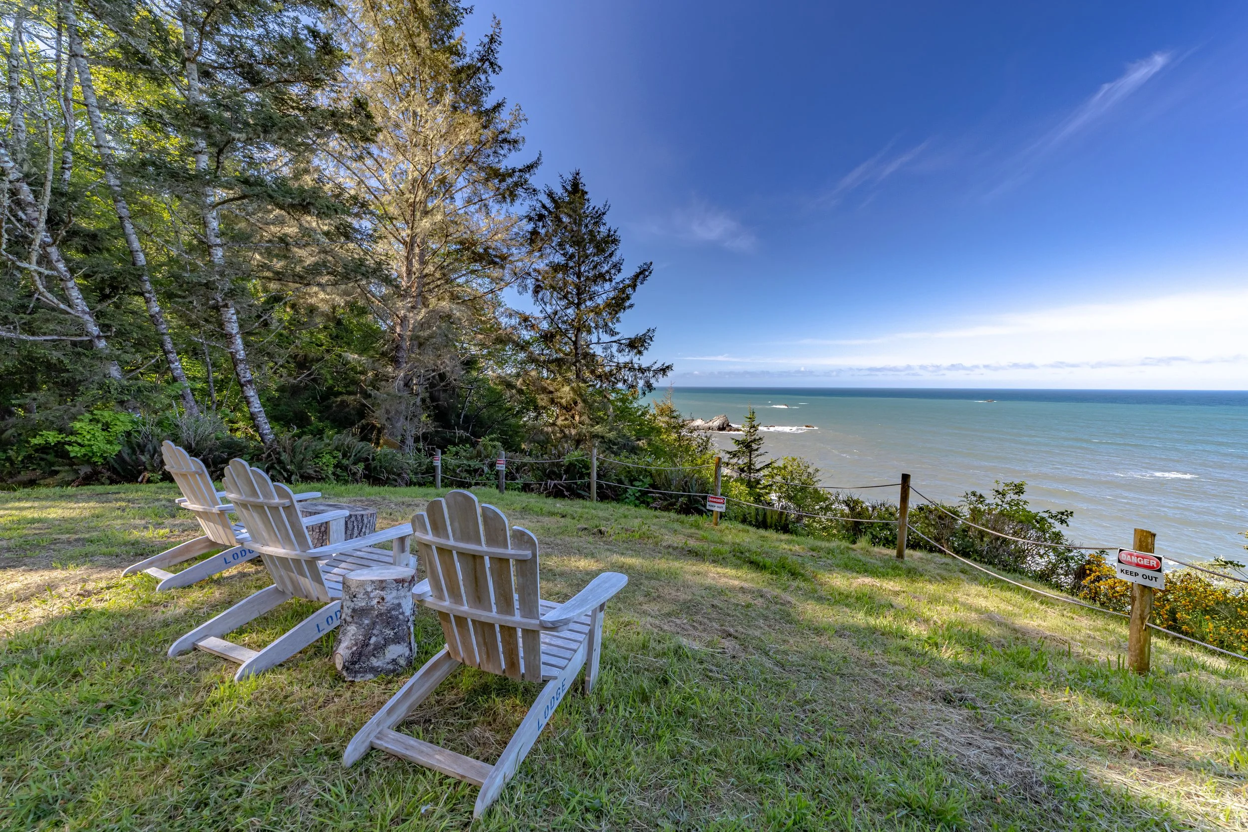 Three wooden Adirondack chairs facing the ocean on a grassy hill near trees with a safety fence and 'Danger Keep Out' signs.