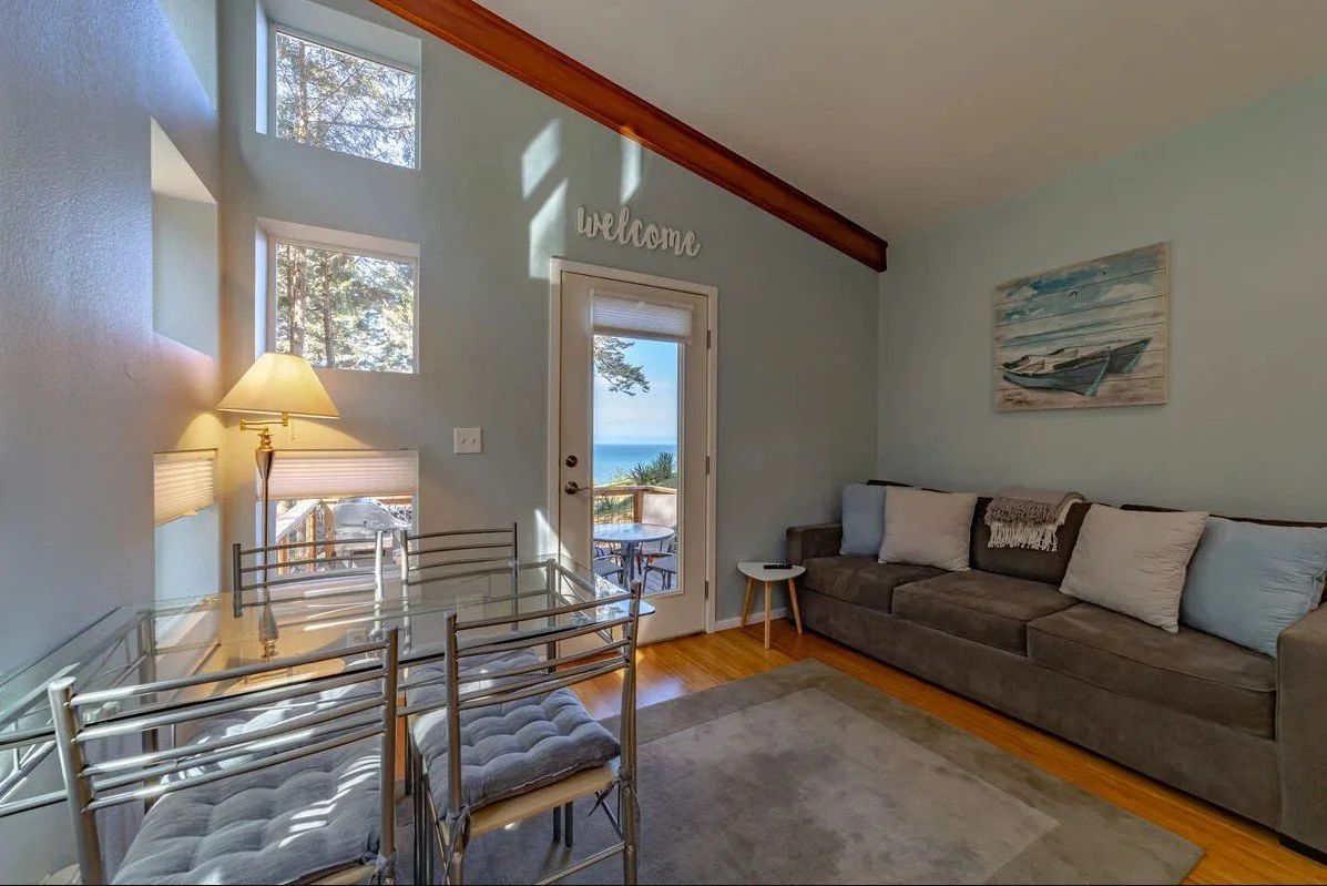 Living room with a large gray sofa, a glass dining table with cushioned chairs, a glass door leading outside, and a view of the ocean through the door and windows. The room has light blue walls, a painting of boats, and a welcome sign above the door.