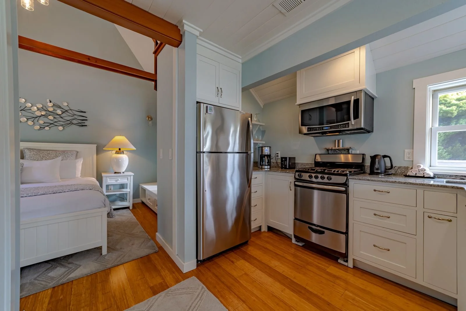 Kitchen with stainless steel refrigerator, stove, microwave, and coffee maker, adjacent to a bedroom with a bed, nightstand, and lamp.