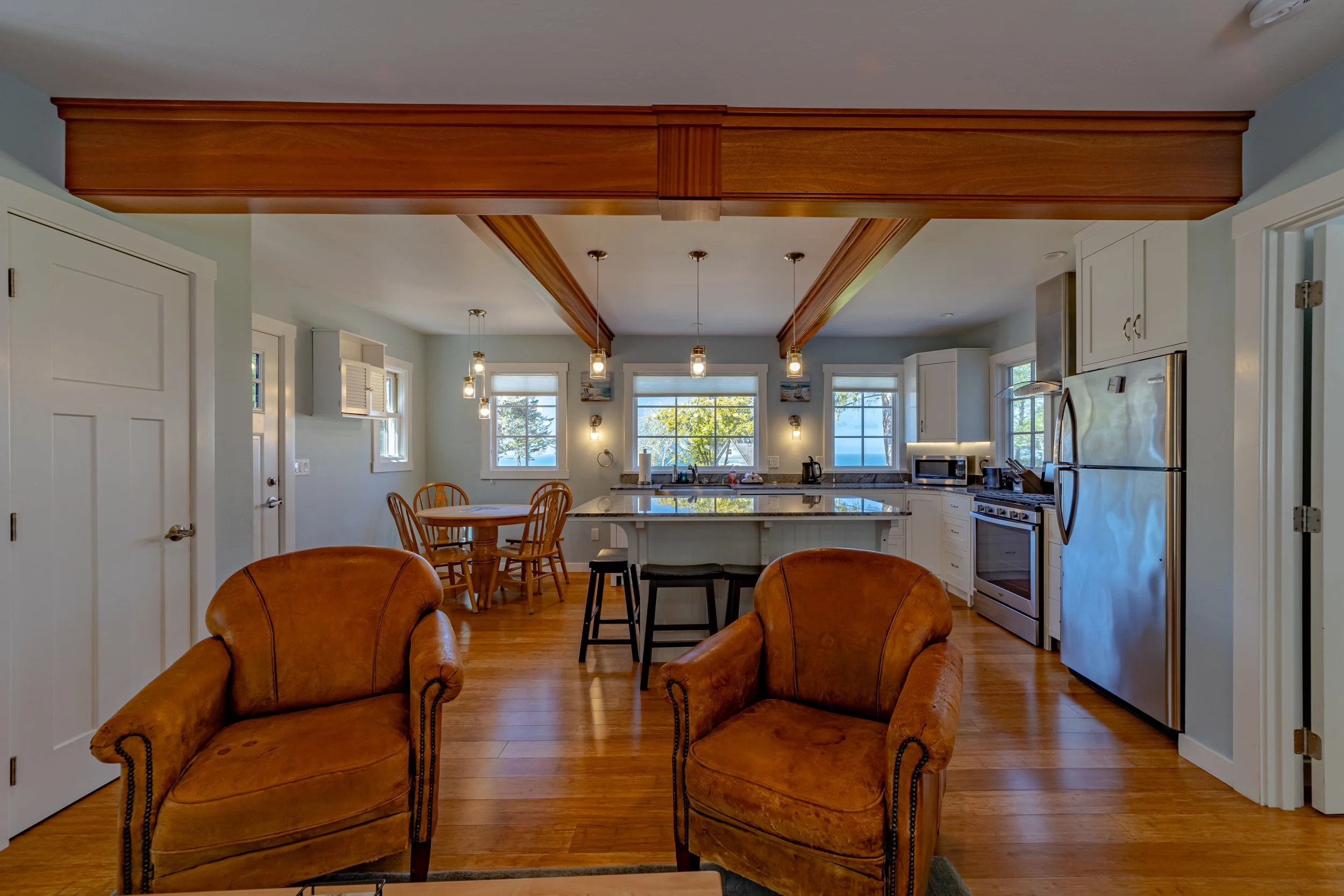 Open-concept kitchen and living area with two brown armchairs, a kitchen island with barstools, white cabinets, stainless steel appliances, wooden floors, and windows with view of trees outside.