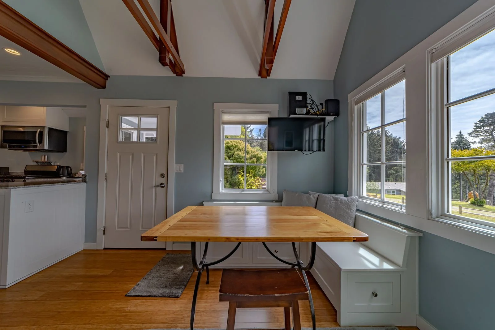 A dining area with a wooden table, bench seating, and windows showing an outdoor view of trees and a yard. A wall-mounted TV is above the bench, and part of a kitchen is visible on the left.