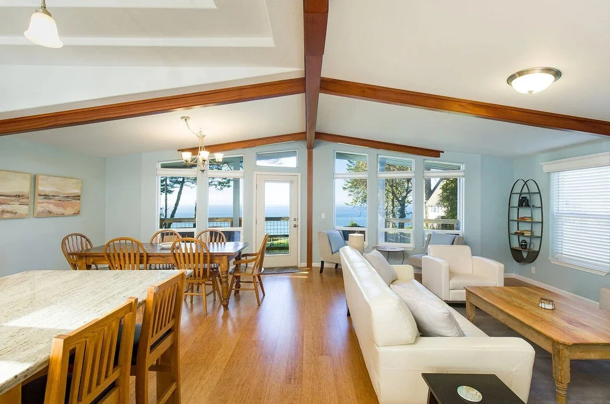 Living room with large windows showing an outdoor view of trees and water, furnished with white sofas, a wooden coffee table, and a dining table with chairs, brightened by natural light and ceiling fixtures.