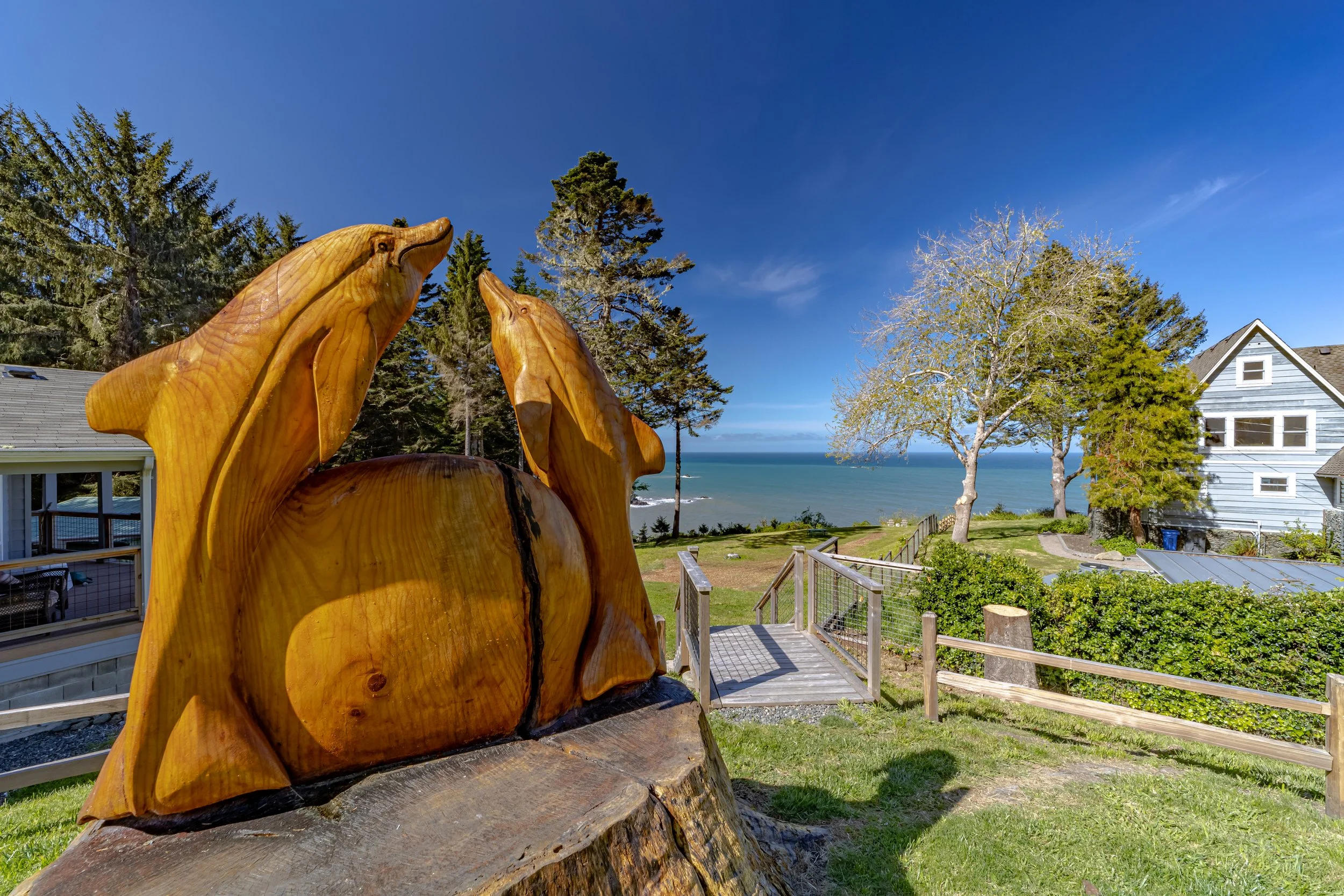 Wooden sculpture of two dolphins near a coastal ocean view with trees, a house, and a clear blue sky.
