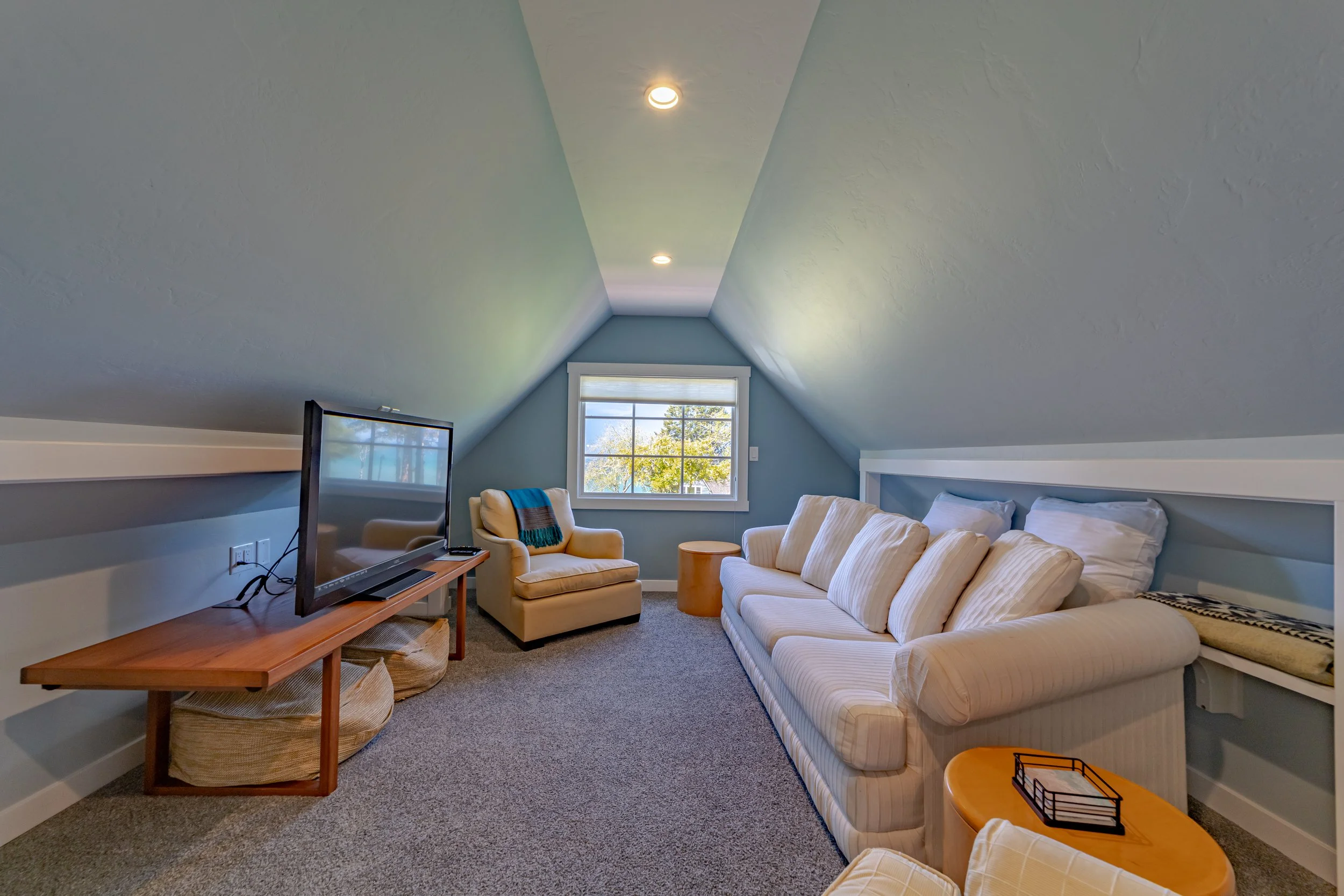 Furnished attic living room with a striped cream sofa, a beige armchair with a blue throw, a wooden side table, a flat-screen TV on a wooden stand, window showing trees outside, recessed ceiling lights, and carpeted floor.