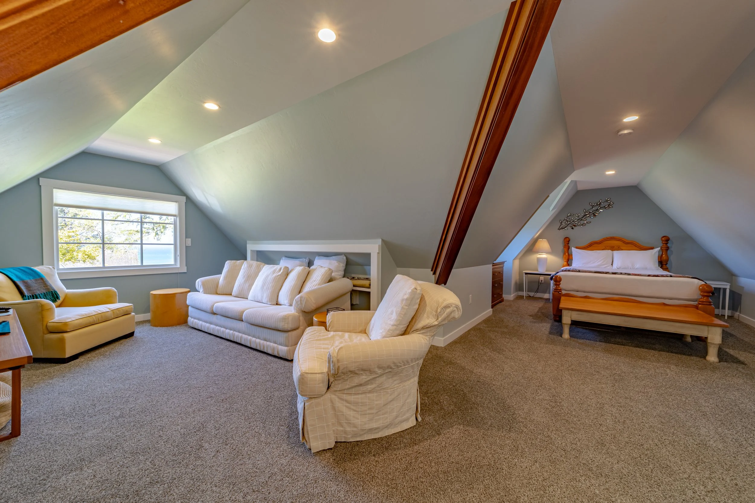 A cozy attic bedroom with sloped ceilings, a sitting area with a yellow armchair and sofa, and a sleeping area with a wooden bed and nightstand, lit by a table lamp.