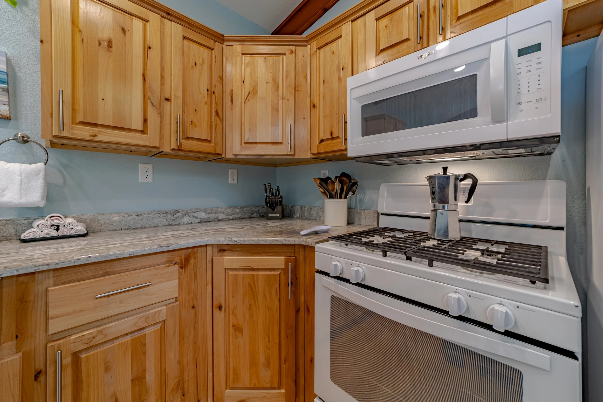 A kitchen with wooden cabinets, granite countertops, a white gas stove with a moka pot on top, a microwave above the stove, and kitchen utensils in a white container.