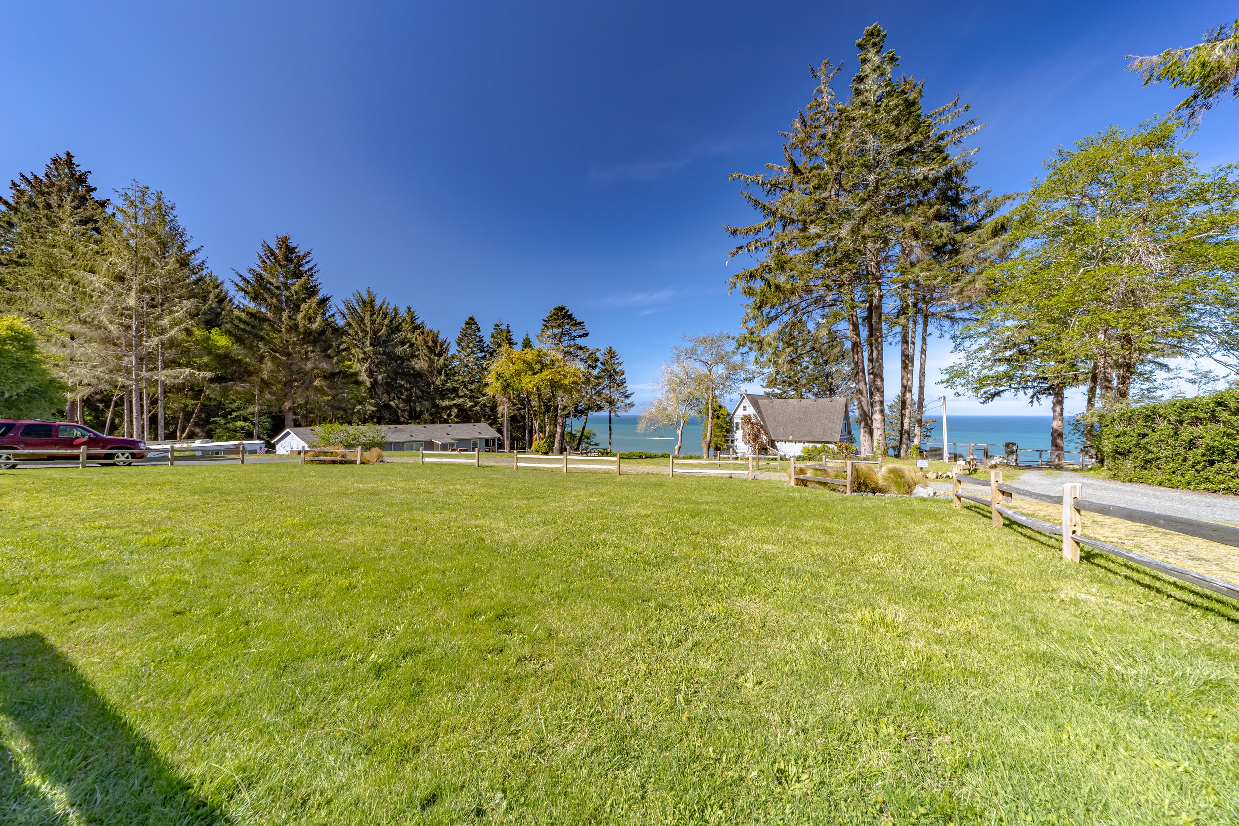 Green lawn with trees, small white houses, a gravel driveway, and a view of the ocean in the background under a clear blue sky.