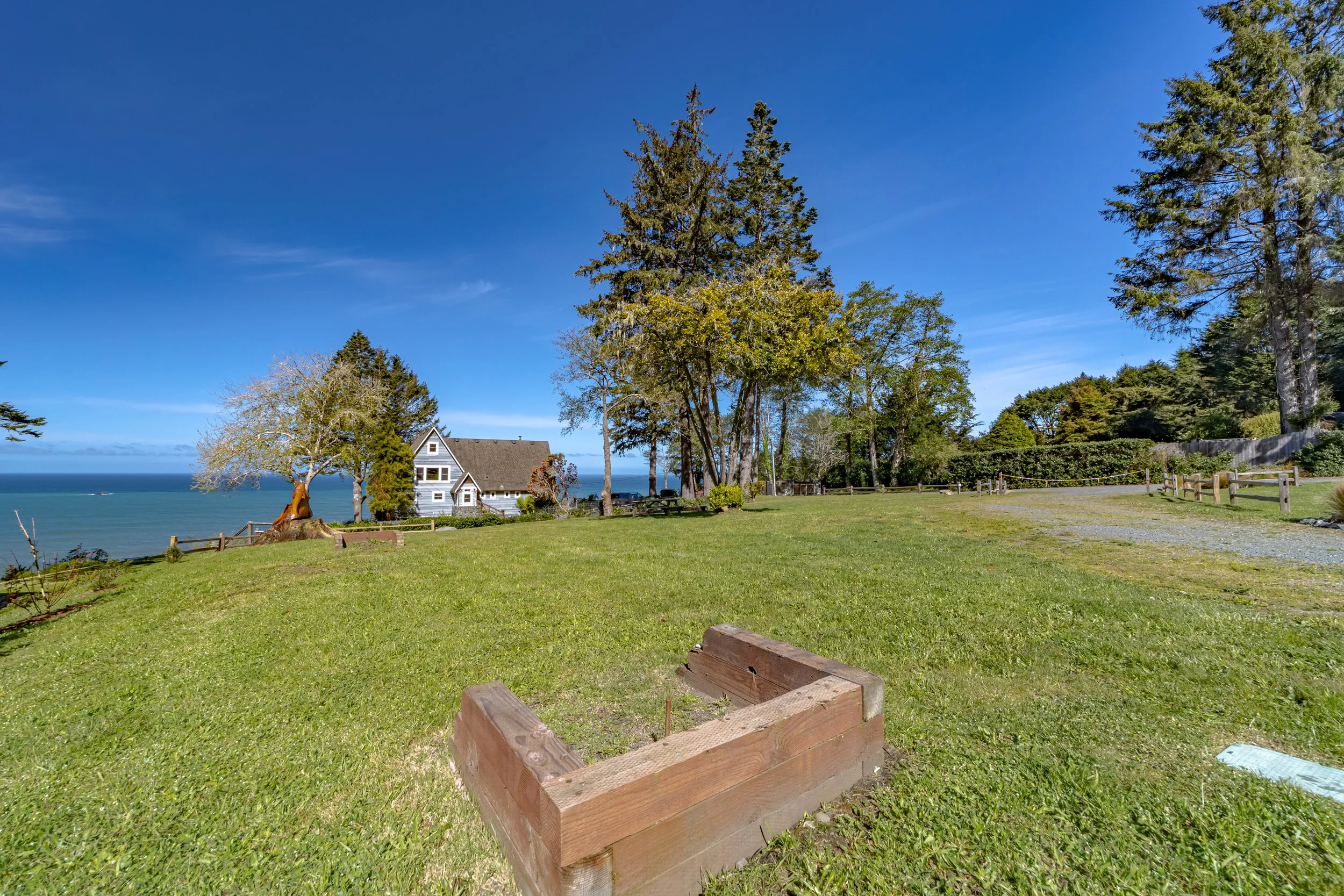 A grassy field with a wooden garden bed in the foreground, trees, a gray house with white trim near the ocean, a dirt path, and a clear blue sky.