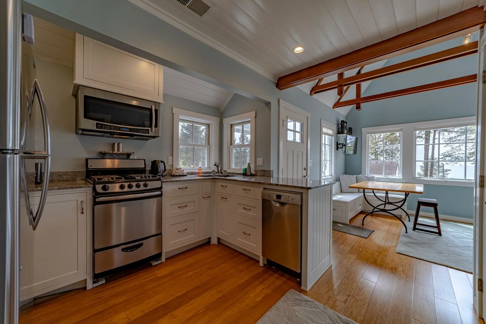 Open-concept kitchen and dining area with white cabinets, stainless steel appliances, light blue walls, and large windows showing trees outside.